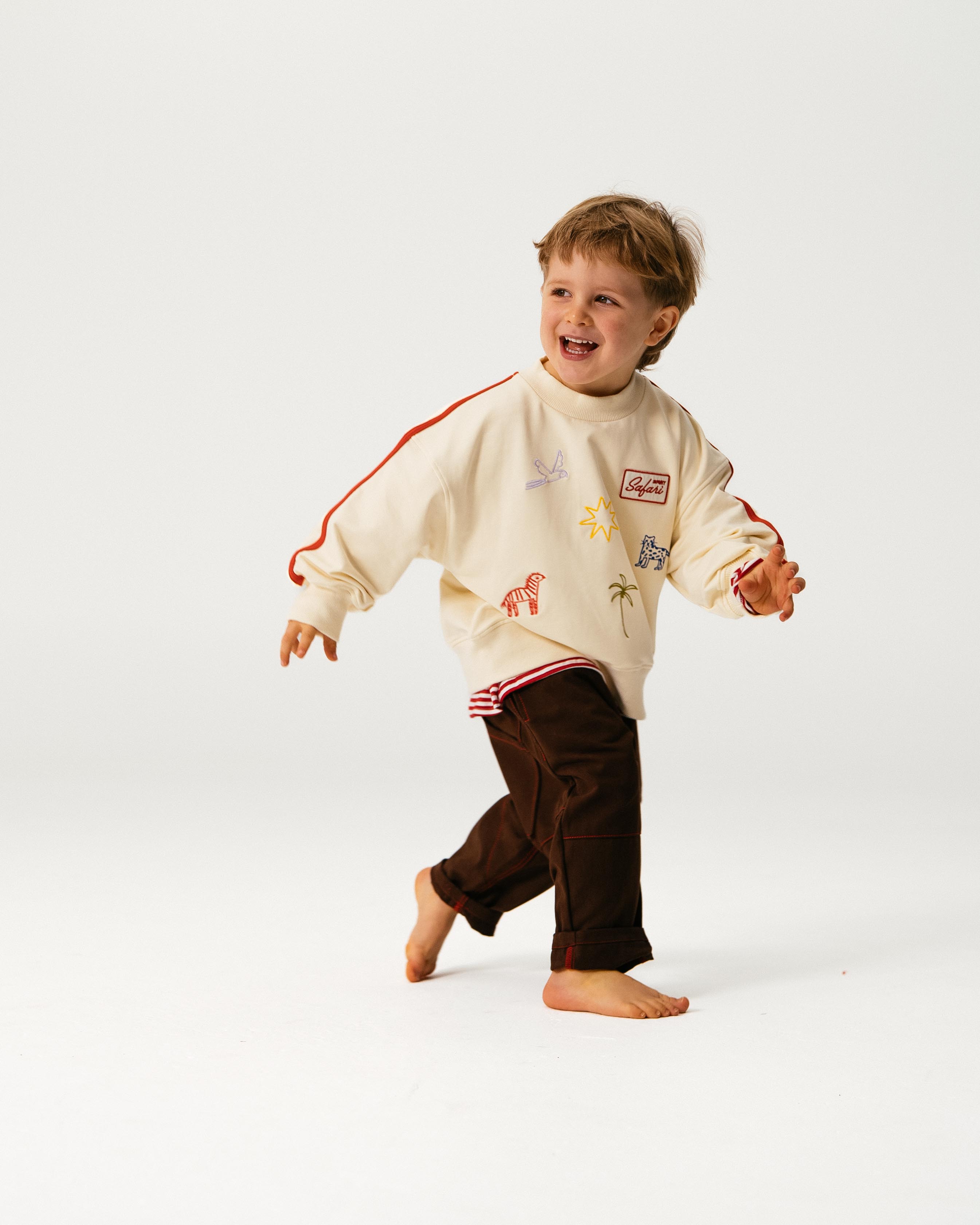 A young child with light brown hair, wearing SONNY LABEL’s cream sweatshirt and Sonny Label - Cargo Pants, smiles while running barefoot against a plain white background.