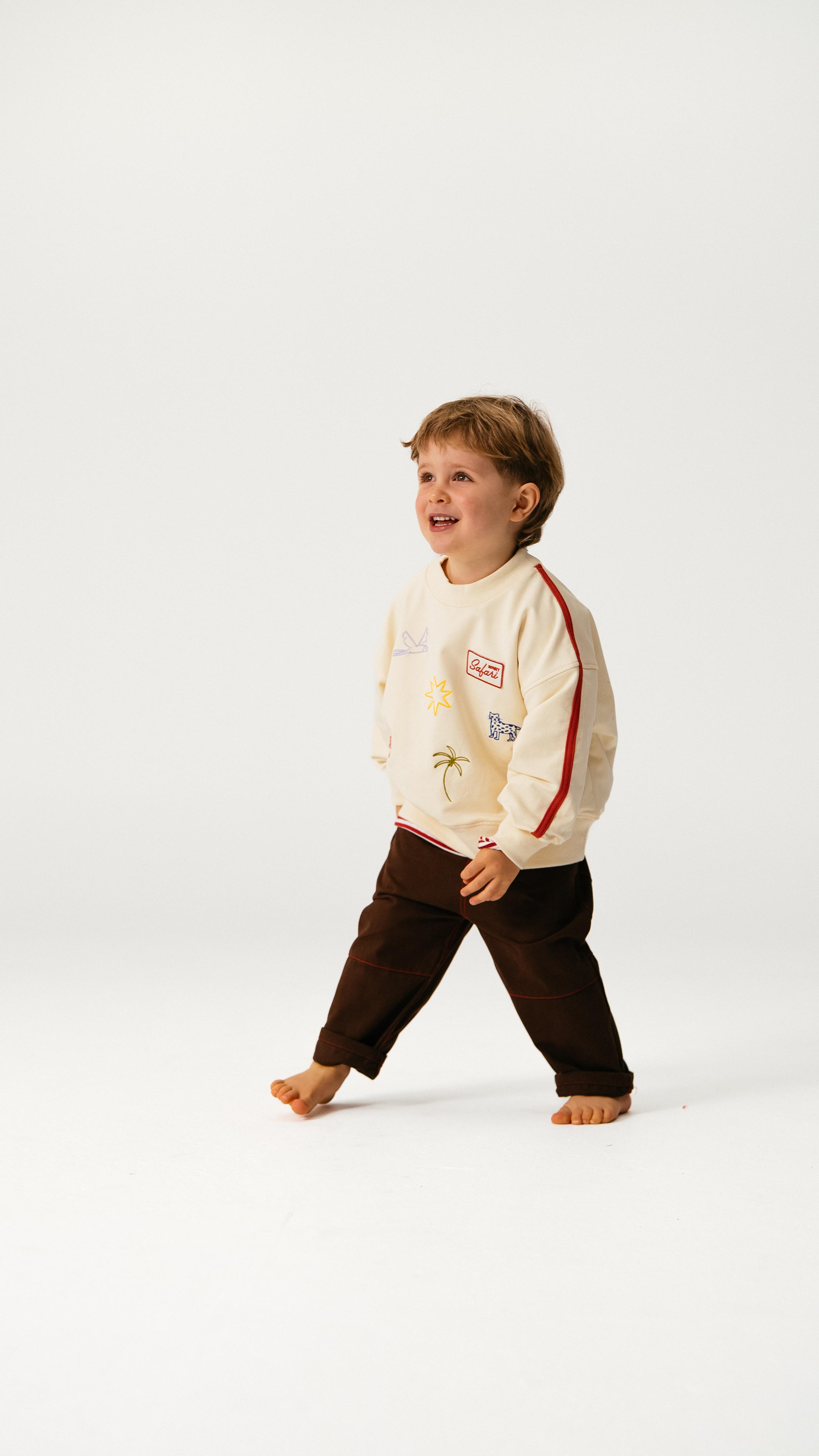 A young child with light brown hair smiles and walks barefoot on a plain white background, wearing a cream sweatshirt and SONNY LABEL Sonny Label - Cargo Pants.