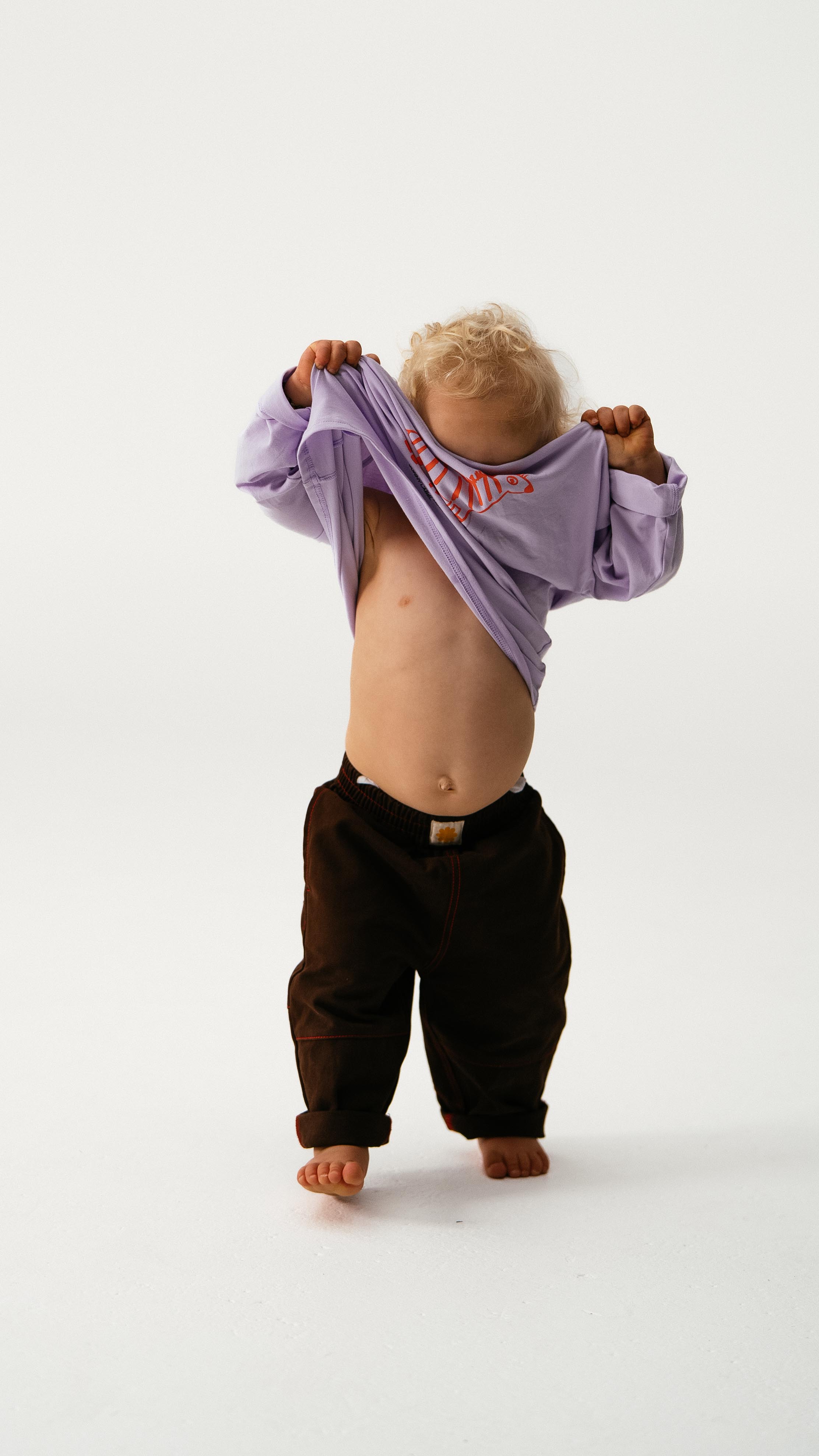 A small child with curly blond hair, barefoot on a white background, lifts a purple shirt over their head and wears dark loose-leg SONNY LABEL Sonny Label - Cargo Pants.
