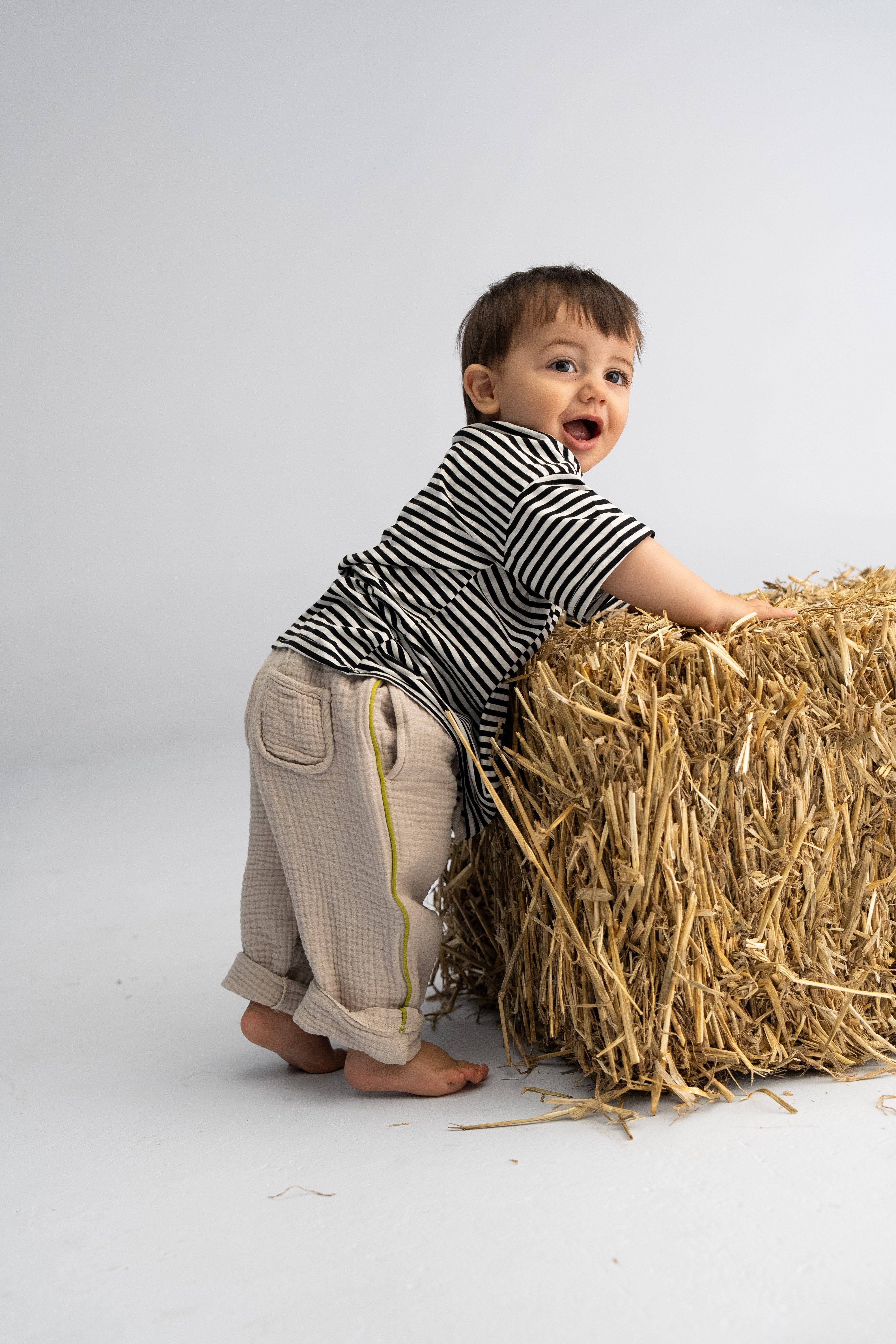 A young child wearing SONNY LABEL's Muslin Pants with Olive Stripe leans against a bale of straw, smiling joyfully. The light, neutral background enhances the playful and cheerful mood, encapsulating the essence of sunny spring days.