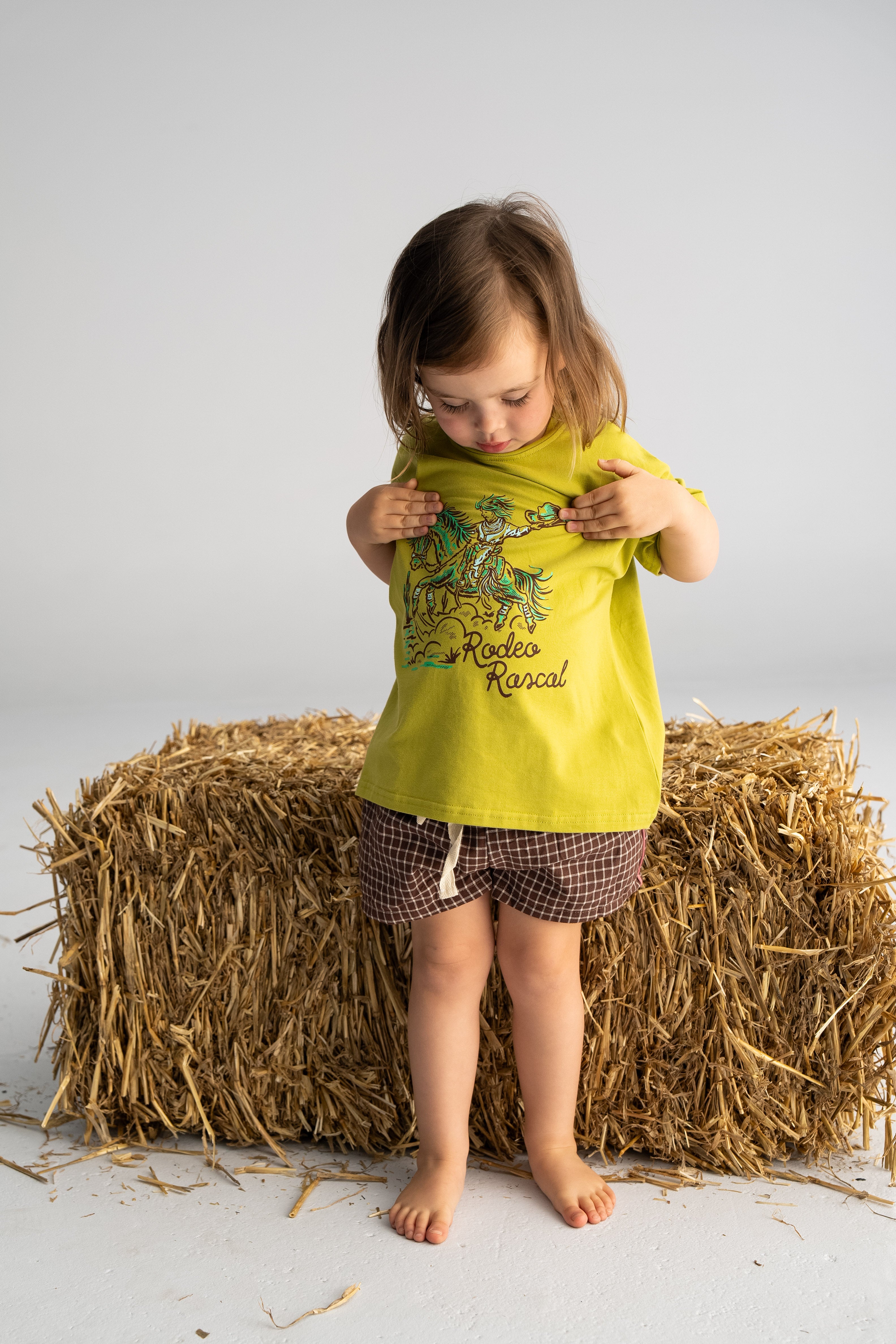 A young child is standing barefoot on a straw bale, sporting the 'Rodeo Rascals' Everyday Tee Olive by SONNY LABEL, paired with checkered shorts. The oversized green tee features a playful Rodeo Rascals illustration, and the child is gazing downward while slightly tugging at the shirt against a plain, light background.