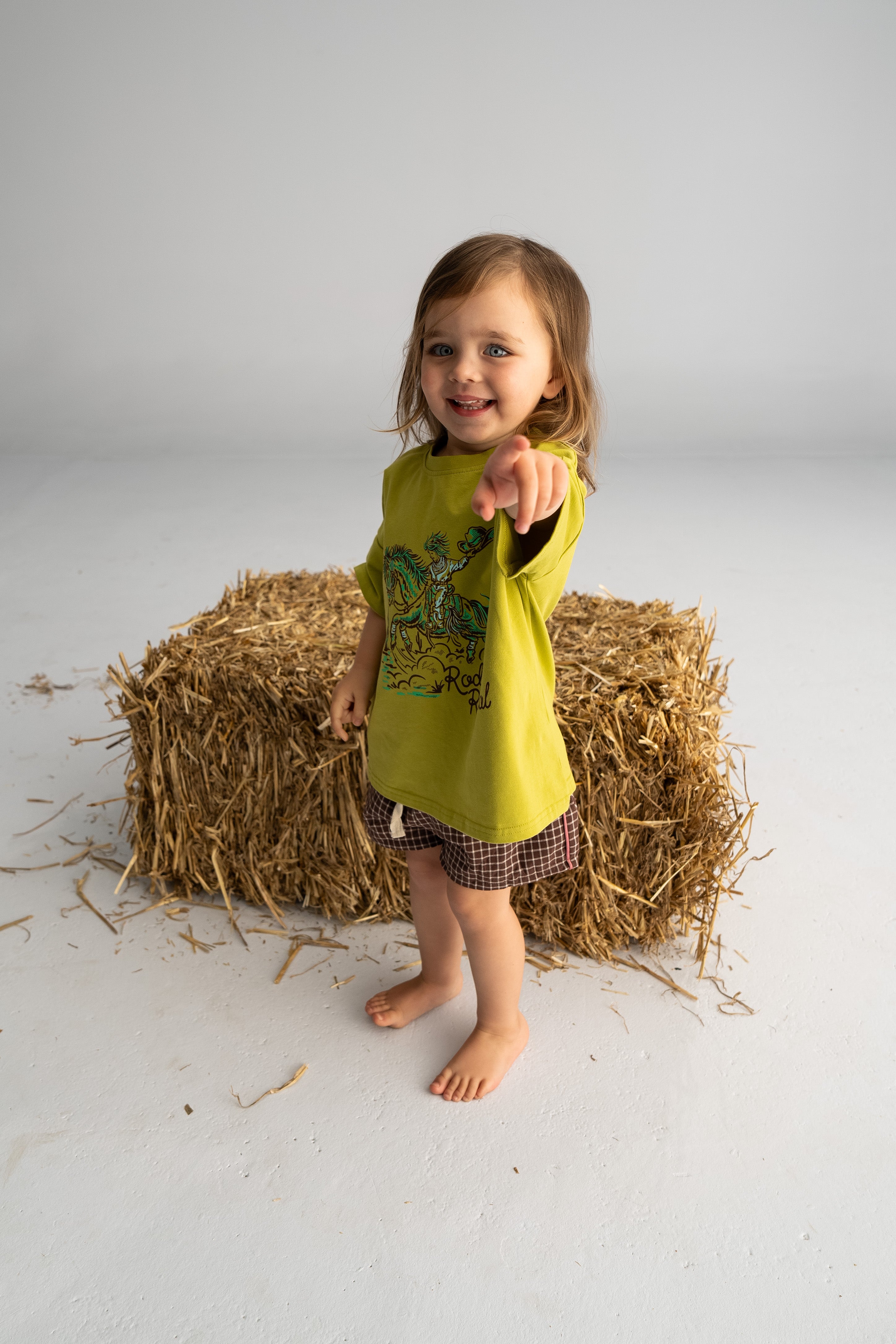 A young child with long hair is smiling and pointing towards the camera, standing barefoot on a light floor. Dressed in 100% cotton, they wear a green t-shirt paired with SONNY LABEL's Check Shorts with Salmon Stripe, while a straw bale in the background adds a country western touch.