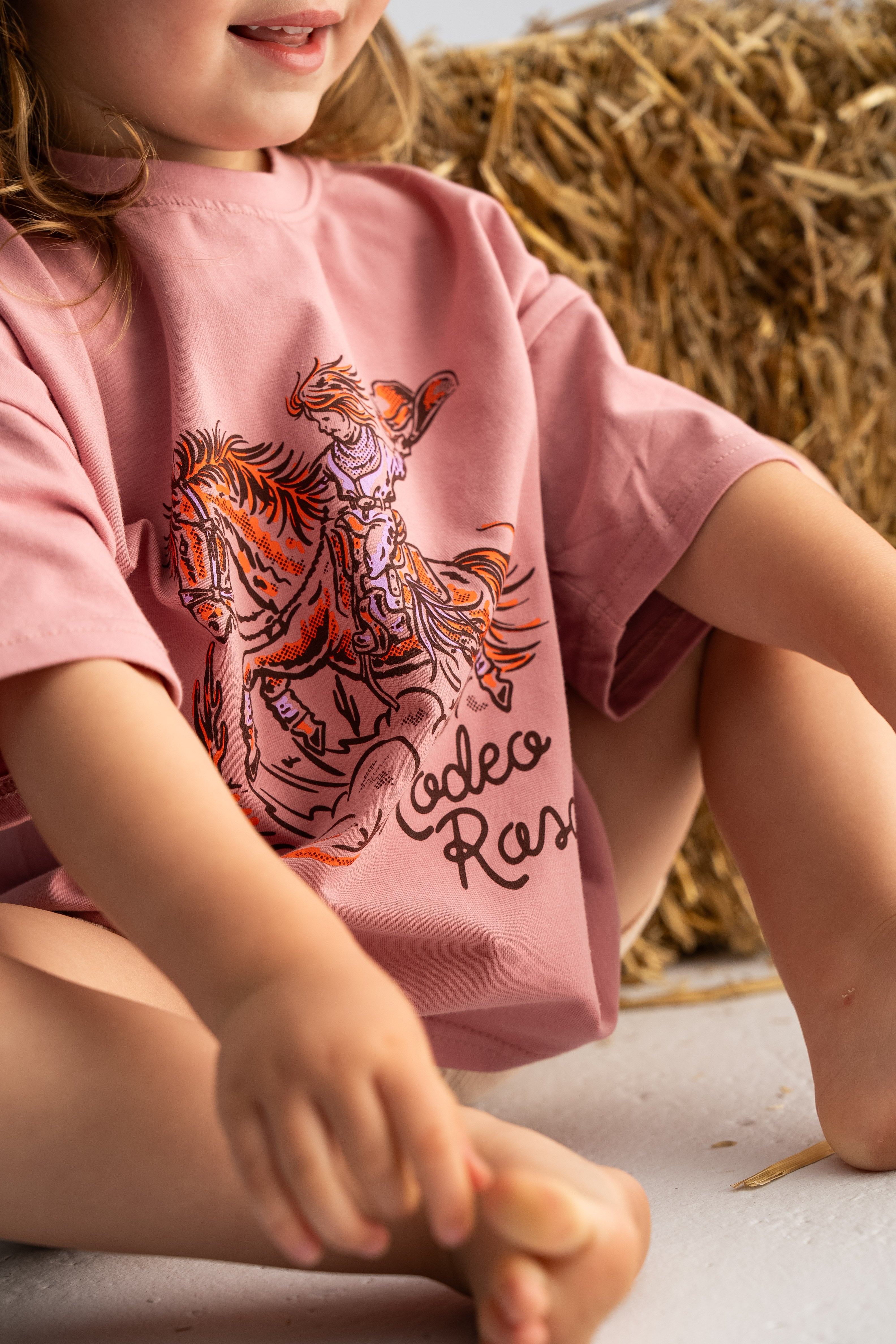 A child sits on the floor wearing a cozy oversized tee from SONNY LABEL's 'Rodeo Rascals' collection, featuring a cowboy illustration and the word "rodeo." The salmon-colored shirt is crafted from soft cotton spandex. They are barefoot with hay bales in the background.