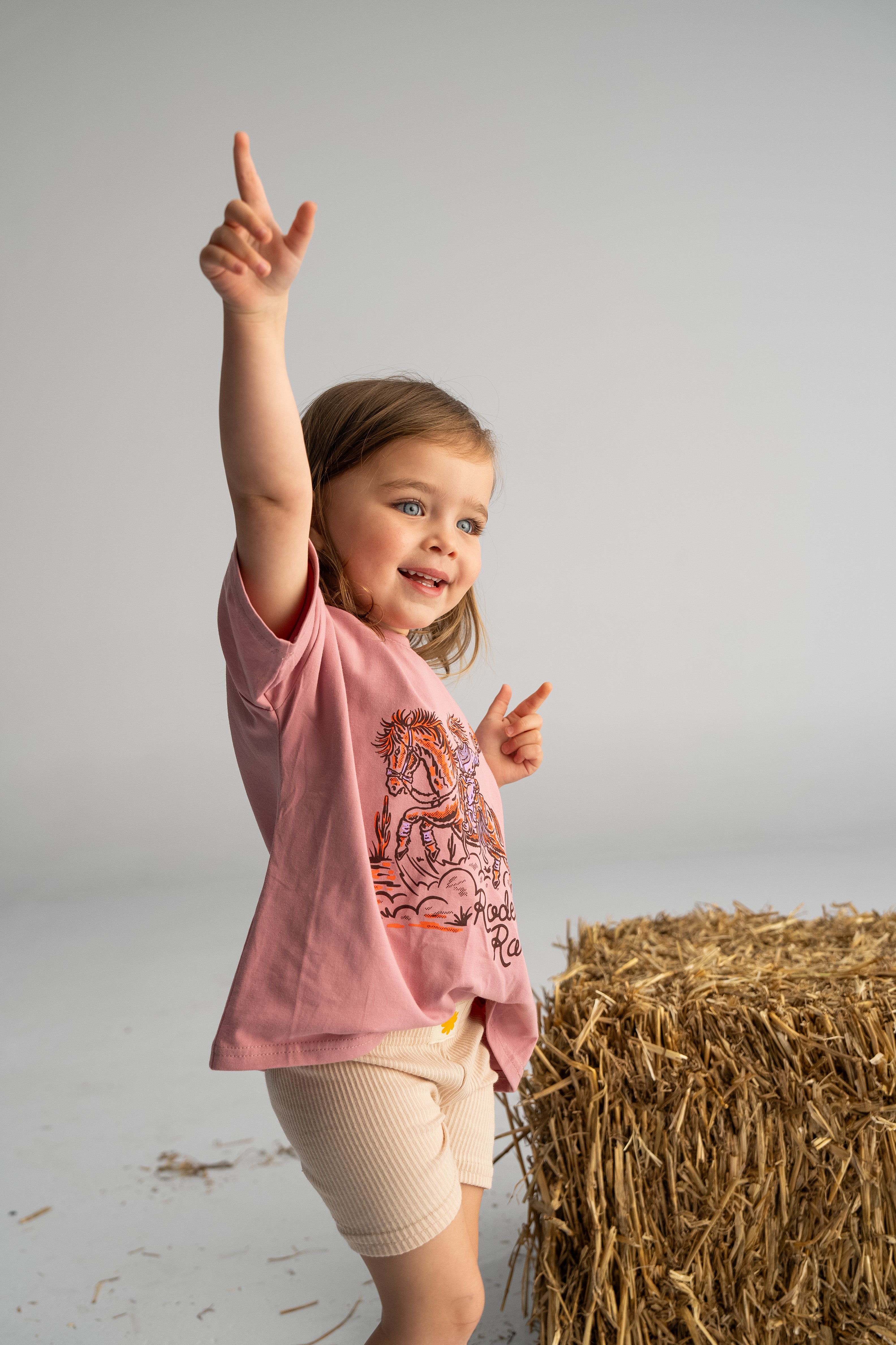 A young child wearing the SONNY LABEL 'Rodeo Rascals' Everyday Tee in Salmon and beige shorts stands next to a hay bale, exuding a cheerful expression that captures the spirit of Rodeo Rascals against a plain, light-colored background.