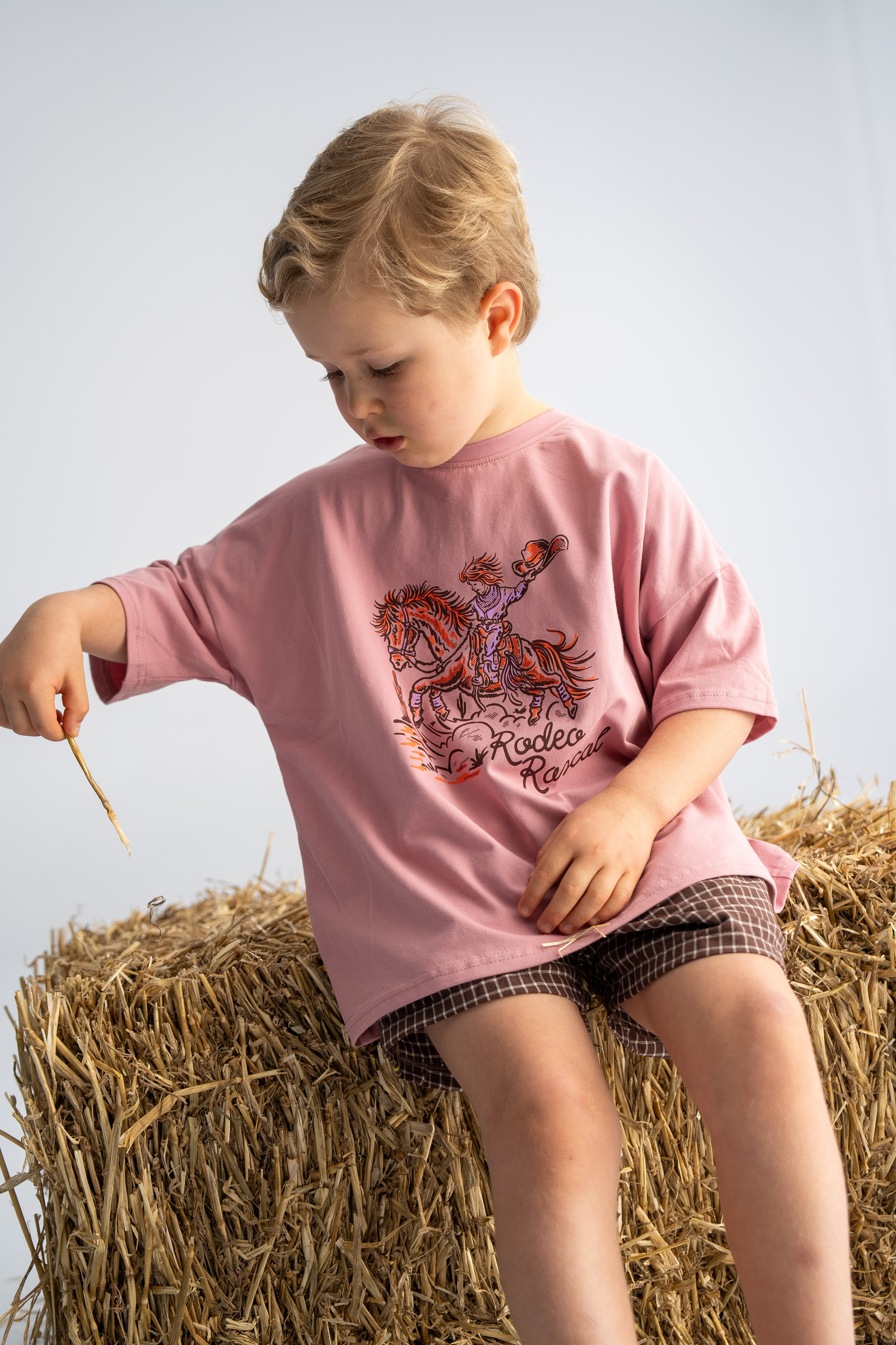 A young child wearing the 'Rodeo Rascals' Everyday Tee in Salmon from SONNY LABEL sits on a bale of hay, holding a small stick. The neutral background accentuates the child's thoughtful expression and casual attire.