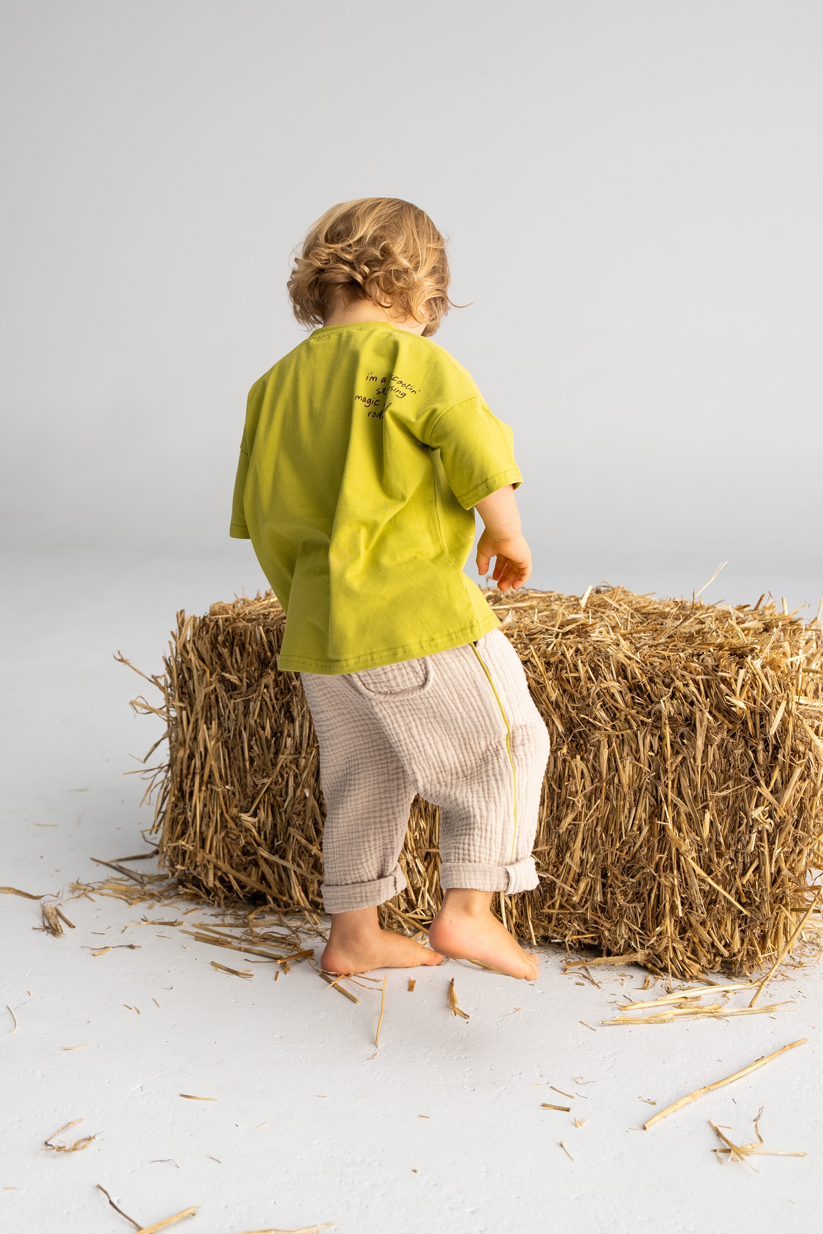 A toddler with curly hair, dressed in a 'Rodeo Rascals' Everyday Tee Olive by SONNY LABEL and beige pants, stands barefoot on a white floor facing a bale of hay. This illustration showcases the child joyfully interacting with the hay against a neutral backdrop.