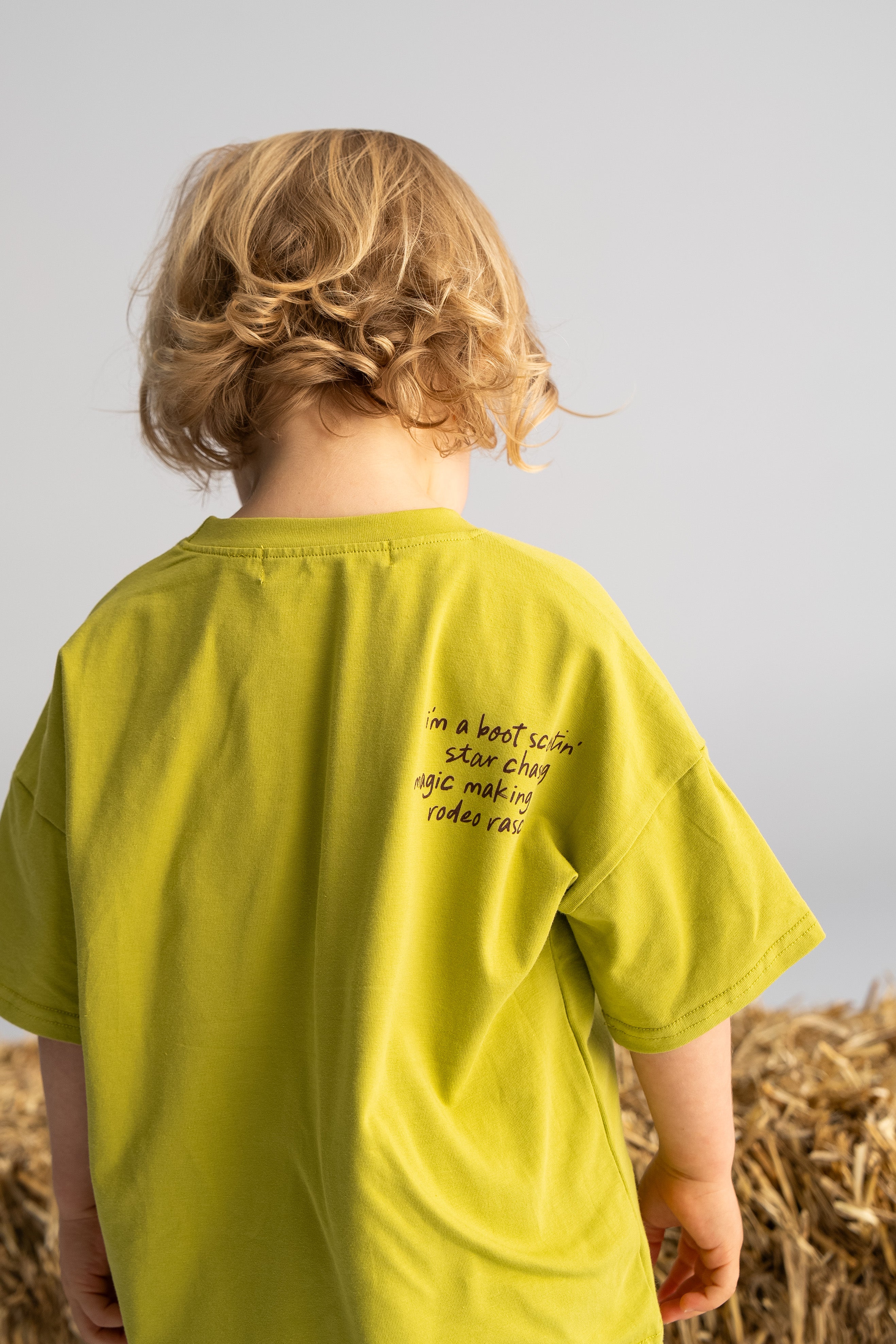 A child with curly blond hair stands facing away, dressed in the SONNY LABEL's 'Rodeo Rascals' Everyday Tee in Olive, showcasing a playful illustration on the back. They are positioned in front of hay bales on a simple gray backdrop.