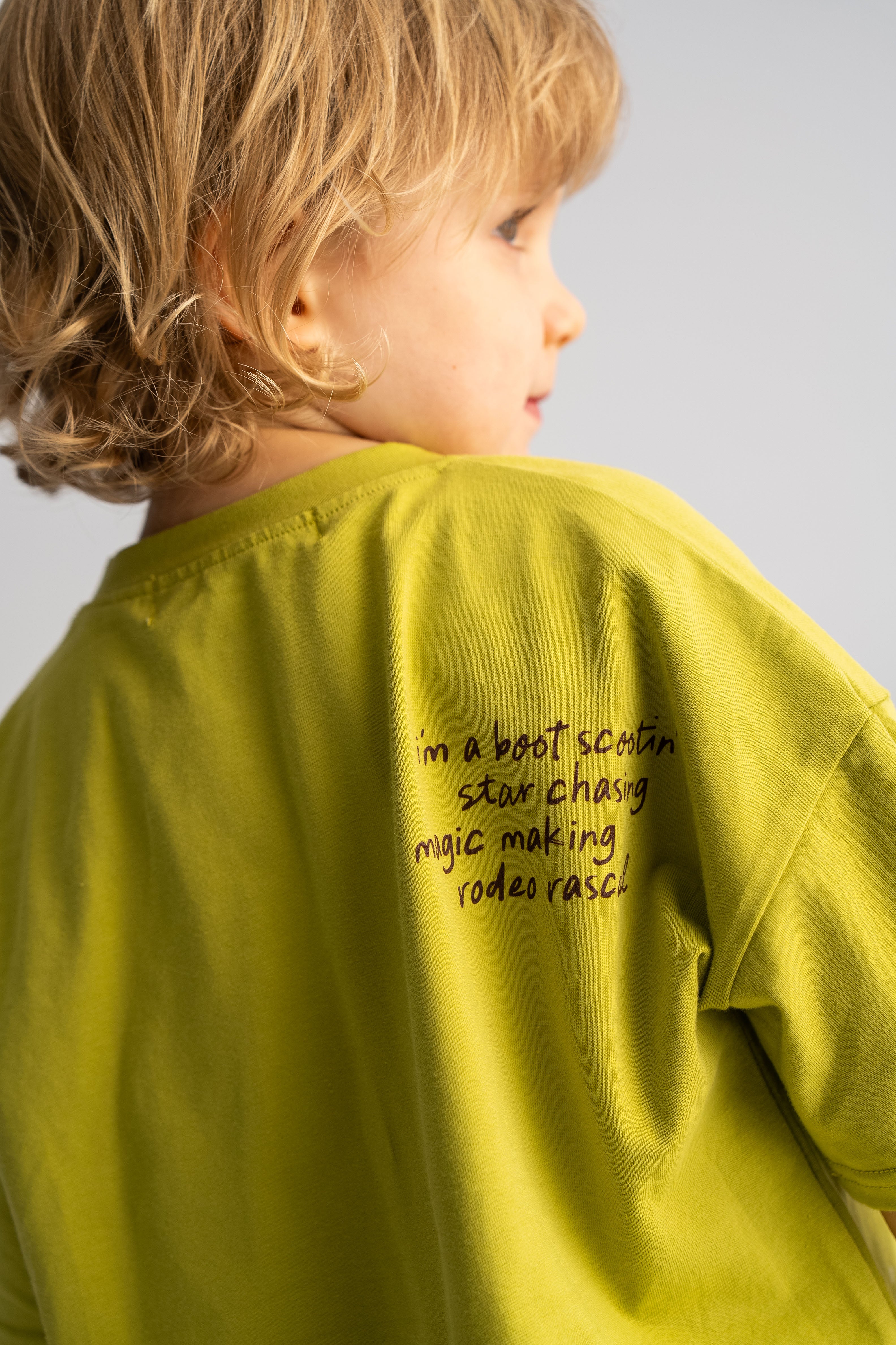 A child with wavy blonde hair wears an oversized olive green everyday tee from SONNY LABEL, featuring the "I'm a boot scootin, star chasing, magic making, Rodeo Rascals" design. The child is turned slightly to the side, gazing off-camera.