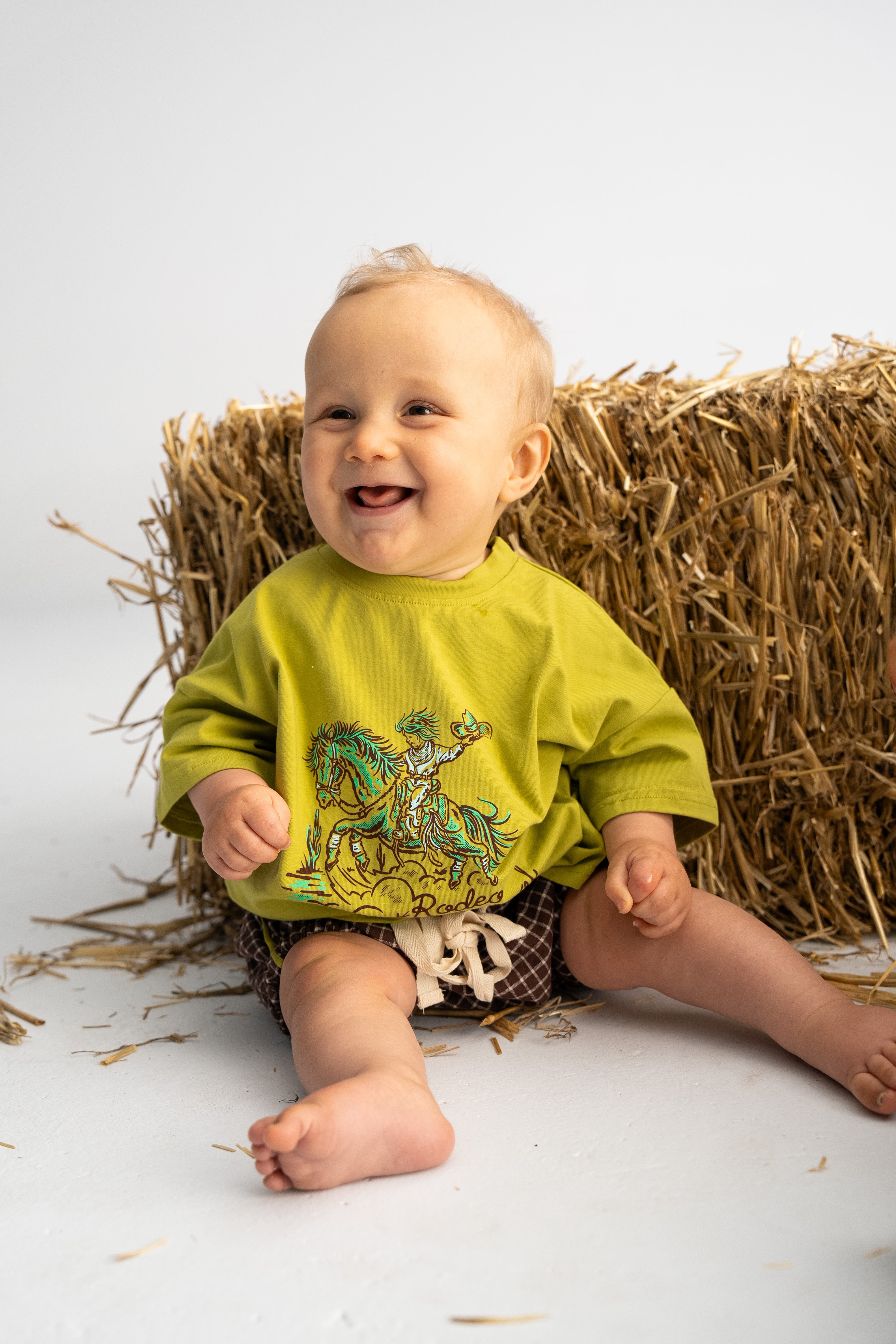 A smiling baby sits on the floor in front of a hay bale, wearing an oversized 'Rodeo Rascals' Everyday Tee in Olive by SONNY LABEL paired with plaid shorts. The background is a plain, light-colored studio setting, highlighting the charming scene.