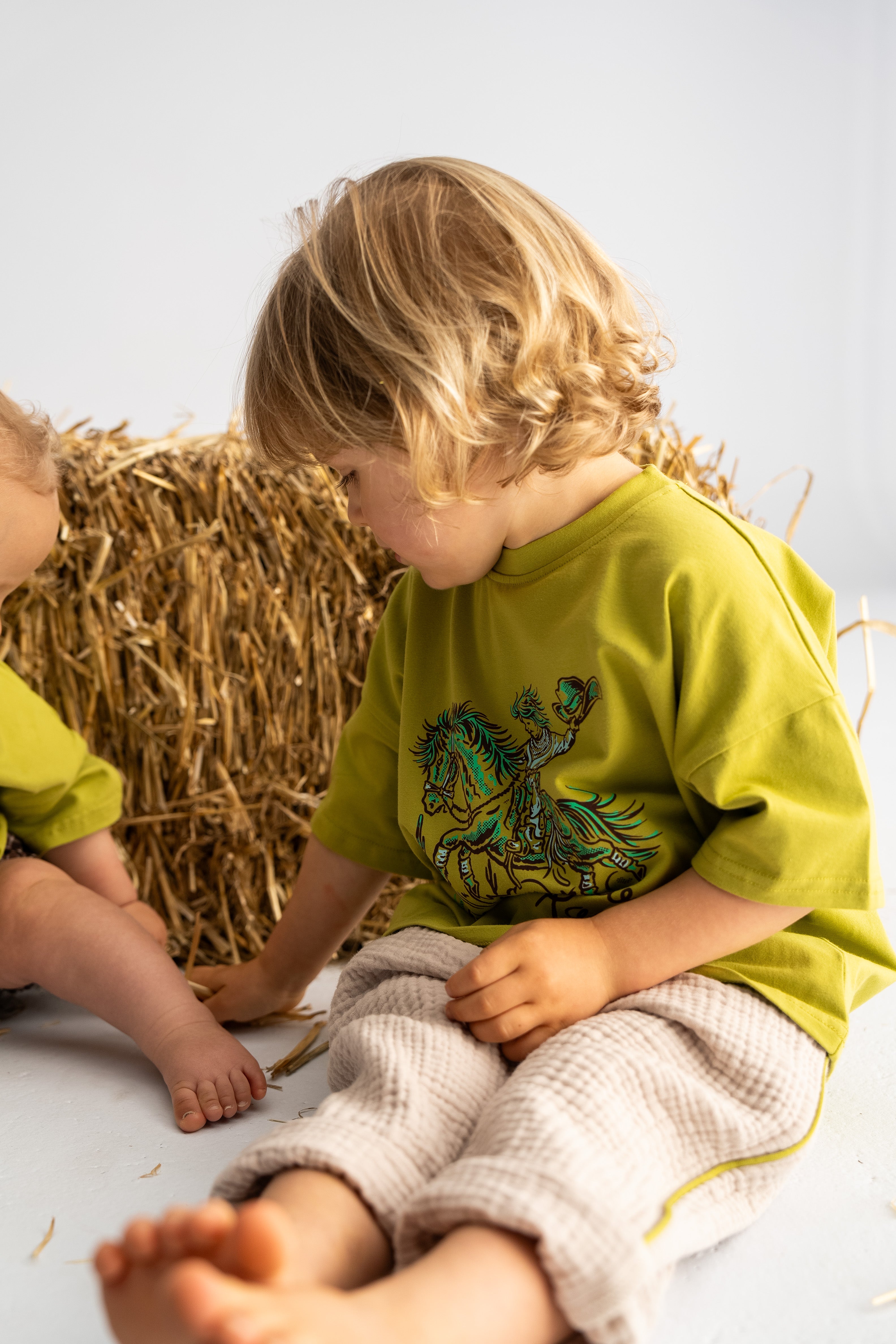 A young child with blonde hair, dressed in a green shirt featuring a horse design, sits on a light floor near a hay bale. Partially visible on the left are the leg and arm of another child, wearing SONNY LABEL's Muslin Pants with Olive Stripe, ideal for spring days. Both children play joyfully with the hay.