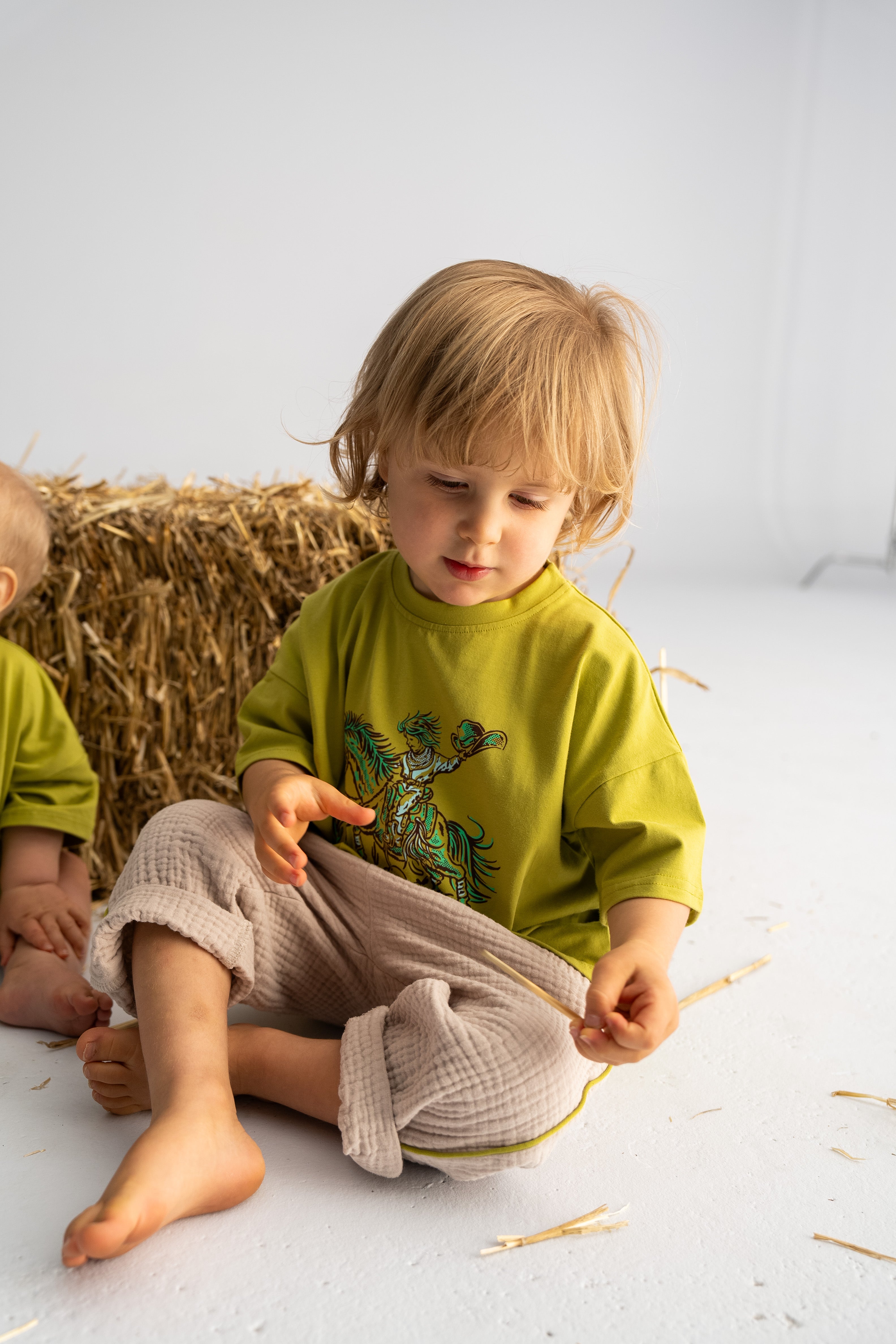 In a bright, minimal room, a young child with blonde hair sits on the floor wearing an oversized 'Rodeo Rascals' Everyday Tee in Olive from SONNY LABEL and beige pants. They play with a piece of straw next to a bale of hay, embodying the playful spirit captured in the Rodeo Rascals illustration.