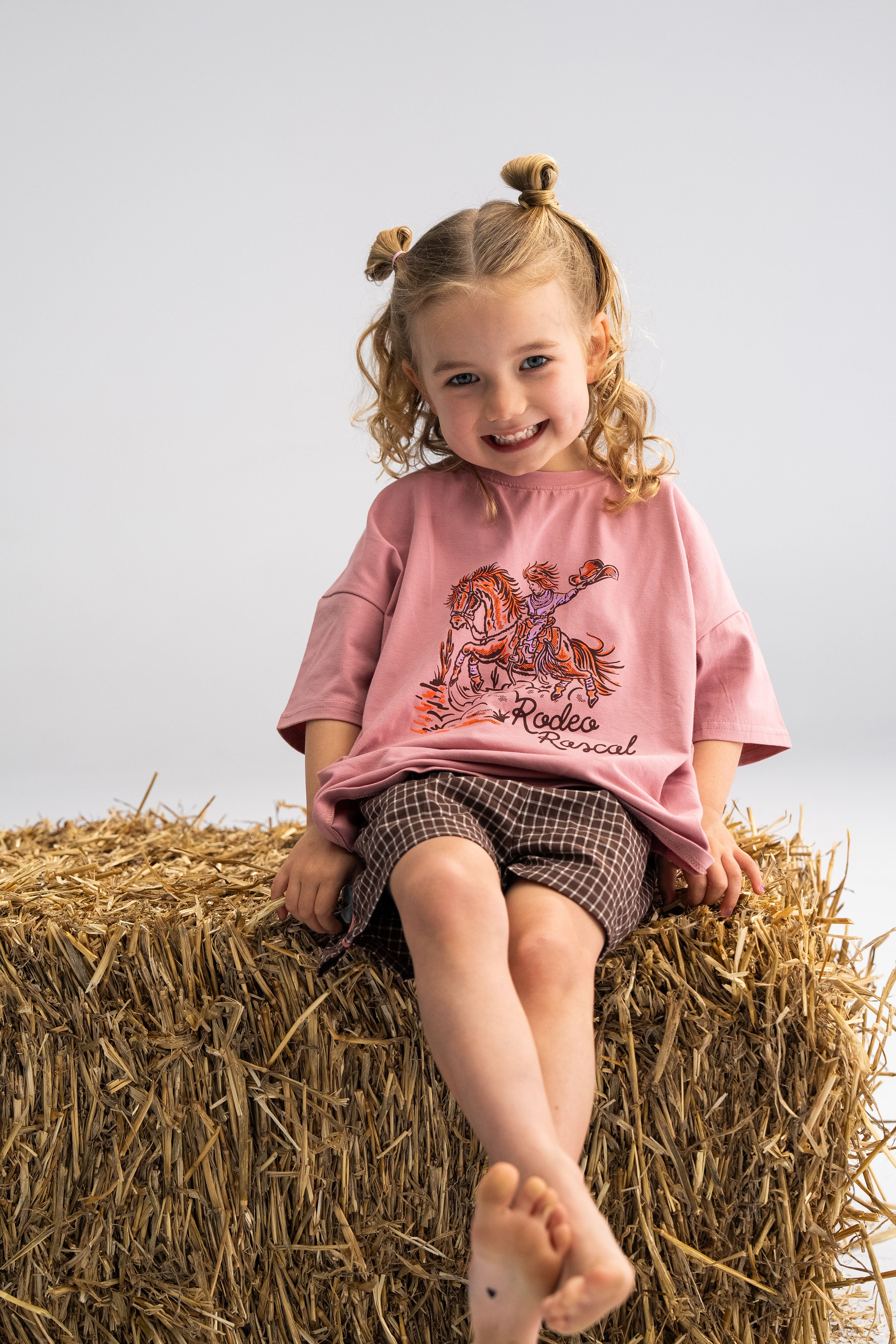 A smiling child with light brown curly hair sits on a hay bale, wearing an oversized 'Rodeo Rascals' Everyday Tee in salmon from SONNY LABEL, paired with brown checkered shorts. Their feet are bare against a simple, light grey background, capturing pure joy and comfort.