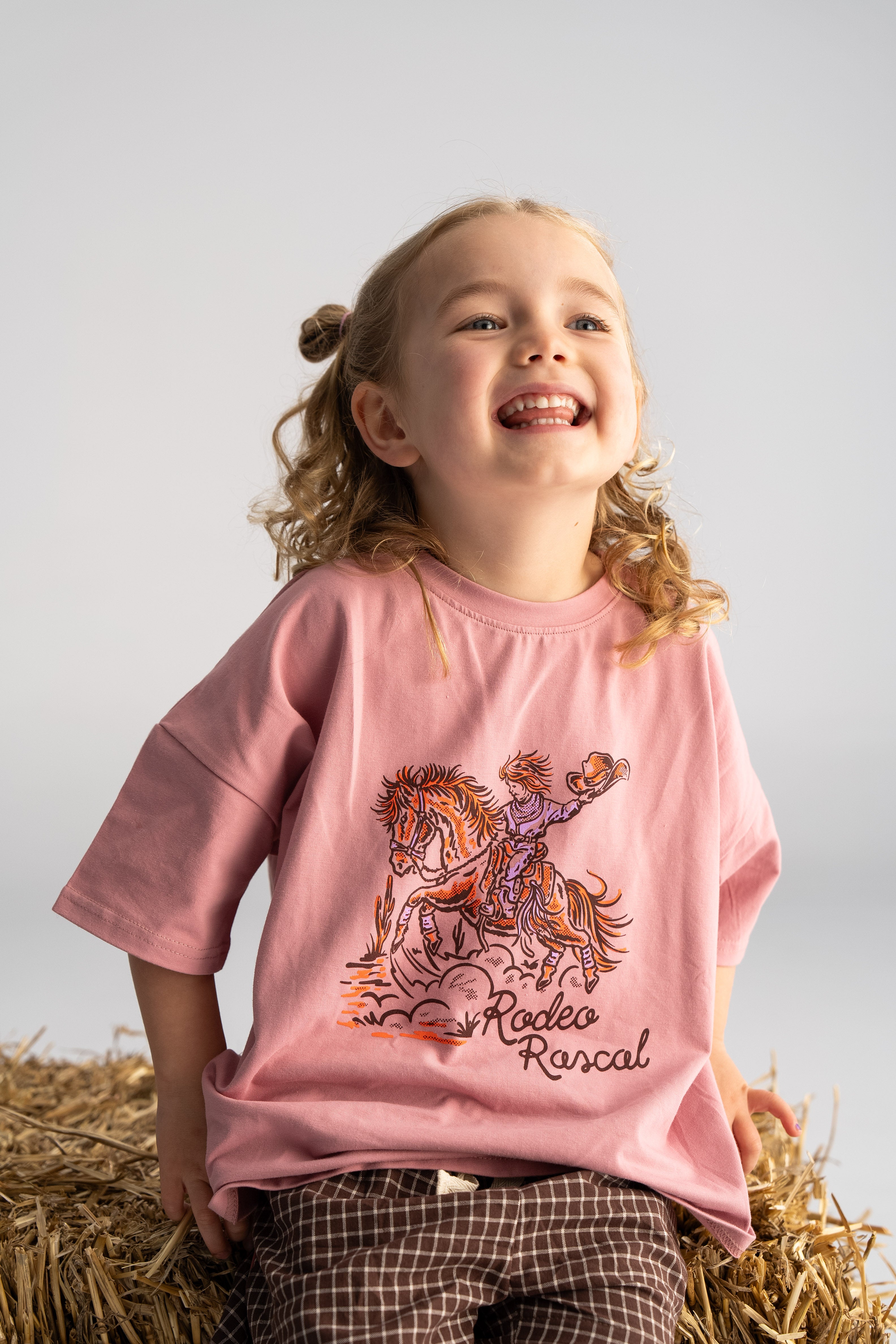 A child with curly hair smiles while sitting on a hay bale, wearing an oversized 'Rodeo Rascals' Everyday Tee in Salmon by SONNY LABEL, adorned with a horse and rider. They have paired it perfectly with cozy cotton spandex plaid pants. The neutral gray backdrop adds charm to the cheerful scene.