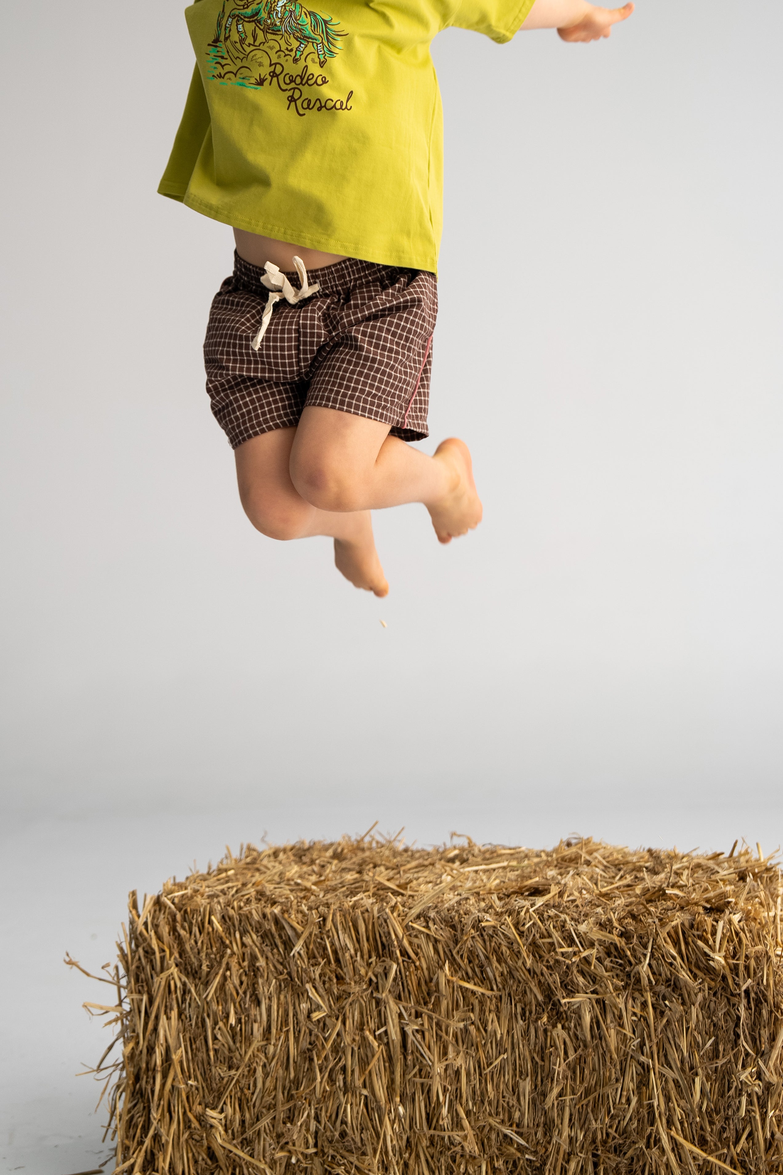 A child wearing SONNY LABEL's Check Shorts with Salmon Stripe and a green shirt leaps above a bale of straw, evoking country western charm in a minimalistic setting. The plain gray background accentuates the scene, with the child's face turned away.