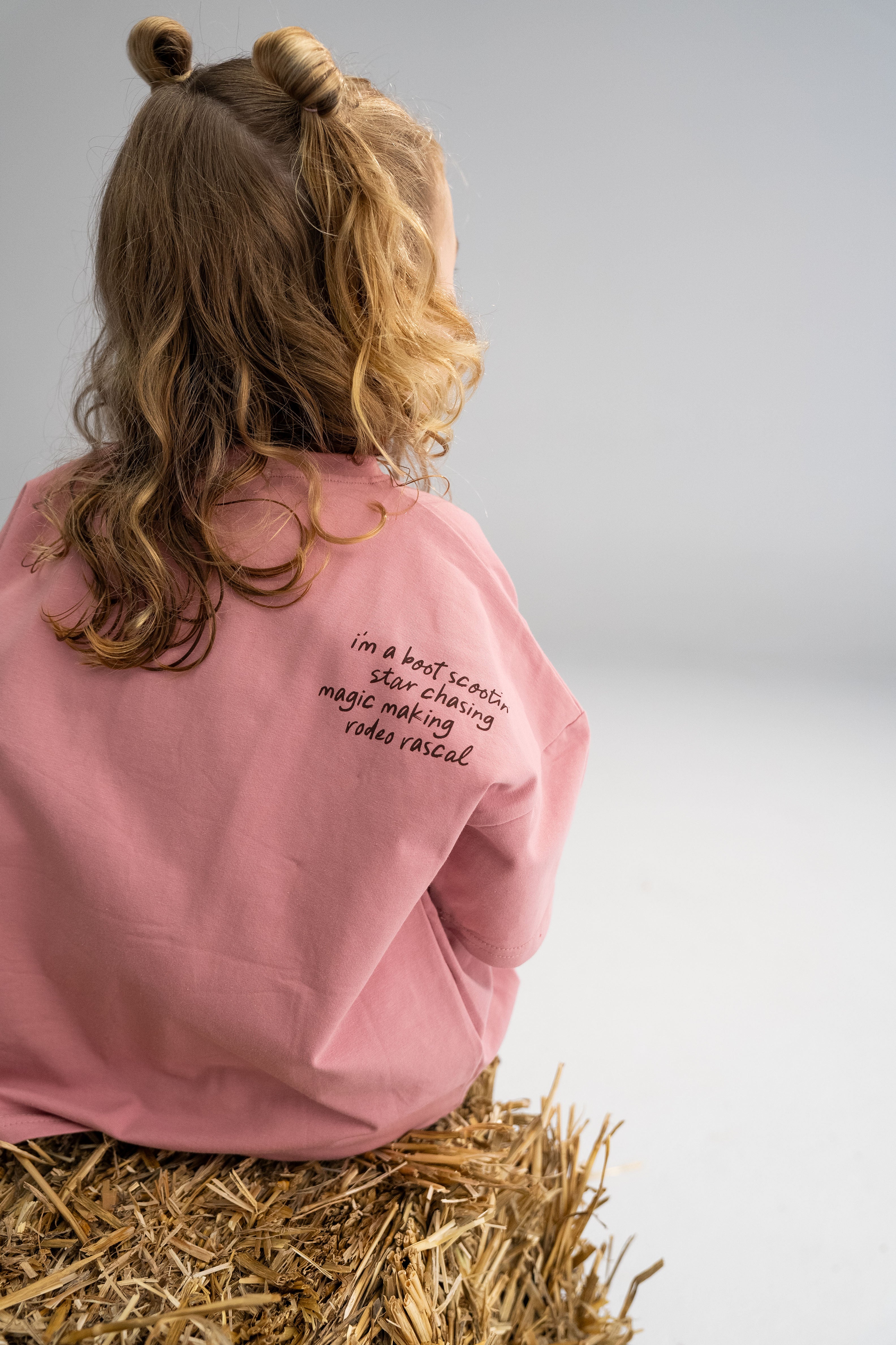 A young child with curly hair sits on a hay bale, wearing an oversized 'Rodeo Rascals' Everyday Tee Salmon by SONNY LABEL. The tee features the text "in a knot scootin, skate chasing, magic making, Rodeo Rascals," and the background is a simple gray.