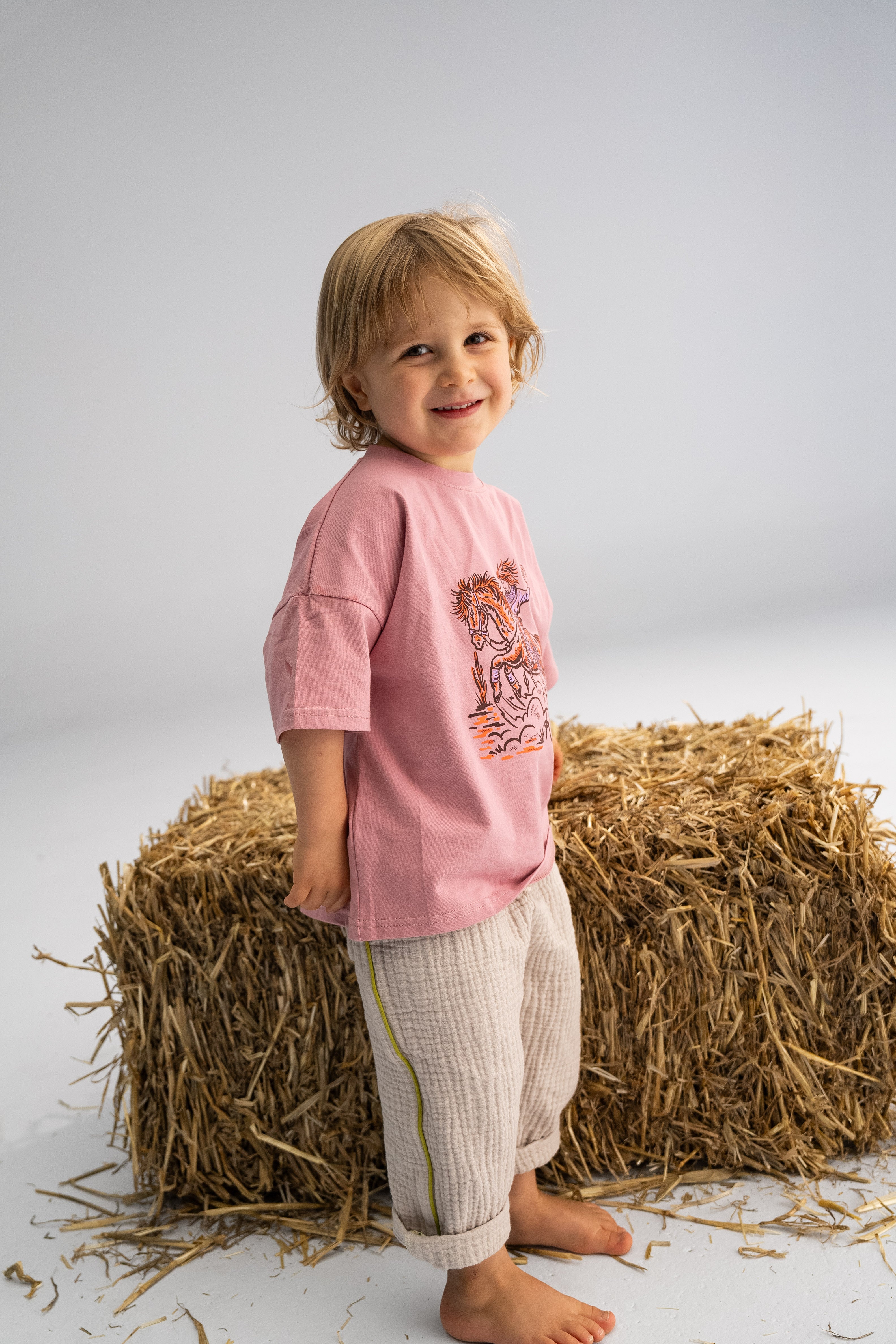 A young child stands smiling barefoot on a white floor next to a hay bale, wearing a pink shirt with an illustrated design and SONNY LABEL's Muslin Pants with Olive Stripe, perfect for the soft embrace of spring days. The background is simple and uncluttered.