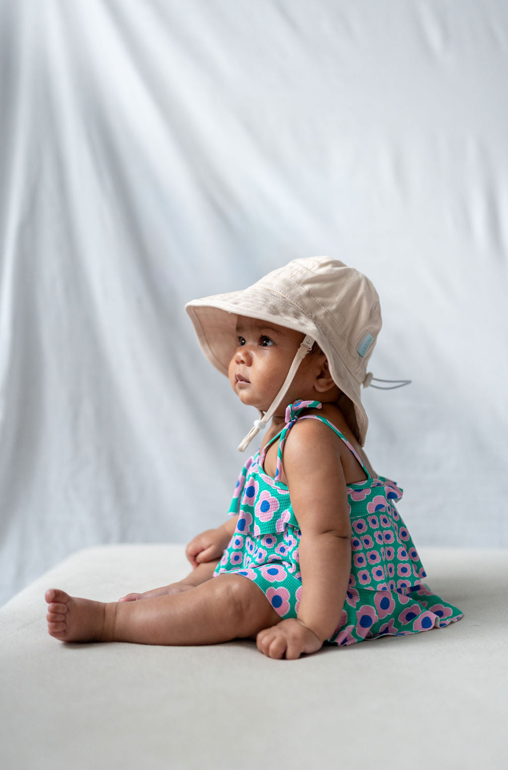 A baby wearing a colorful dress with a geometric pattern and an ACORN KIDS Wide Brim Infant Hat in Vanilla, made of cotton canvas with UPF50+ sun protection, sits on a white surface against a plain white background, looking to the side.