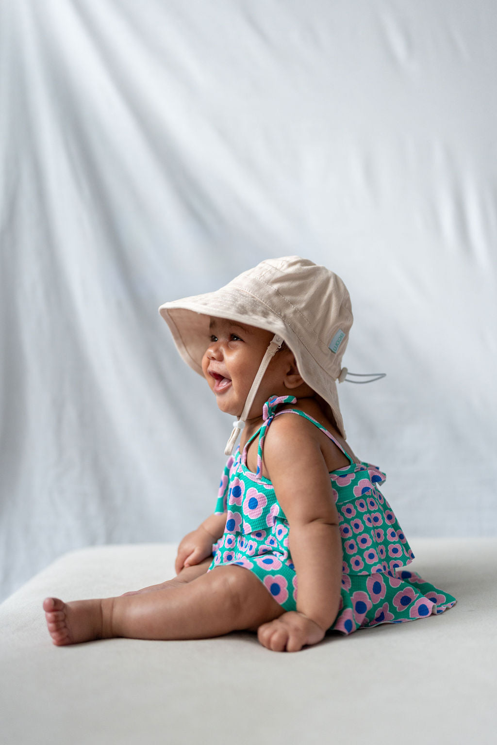 A baby, dressed in a colorful patterned dress and wearing the ACORN KIDS' Wide Brim Infant Hat in Vanilla, which offers UPF50+ sun protection, sits and smiles with a white backdrop behind them. The baby appears happy and engaged, looking slightly to the side. The photo captures a light, joyful moment.