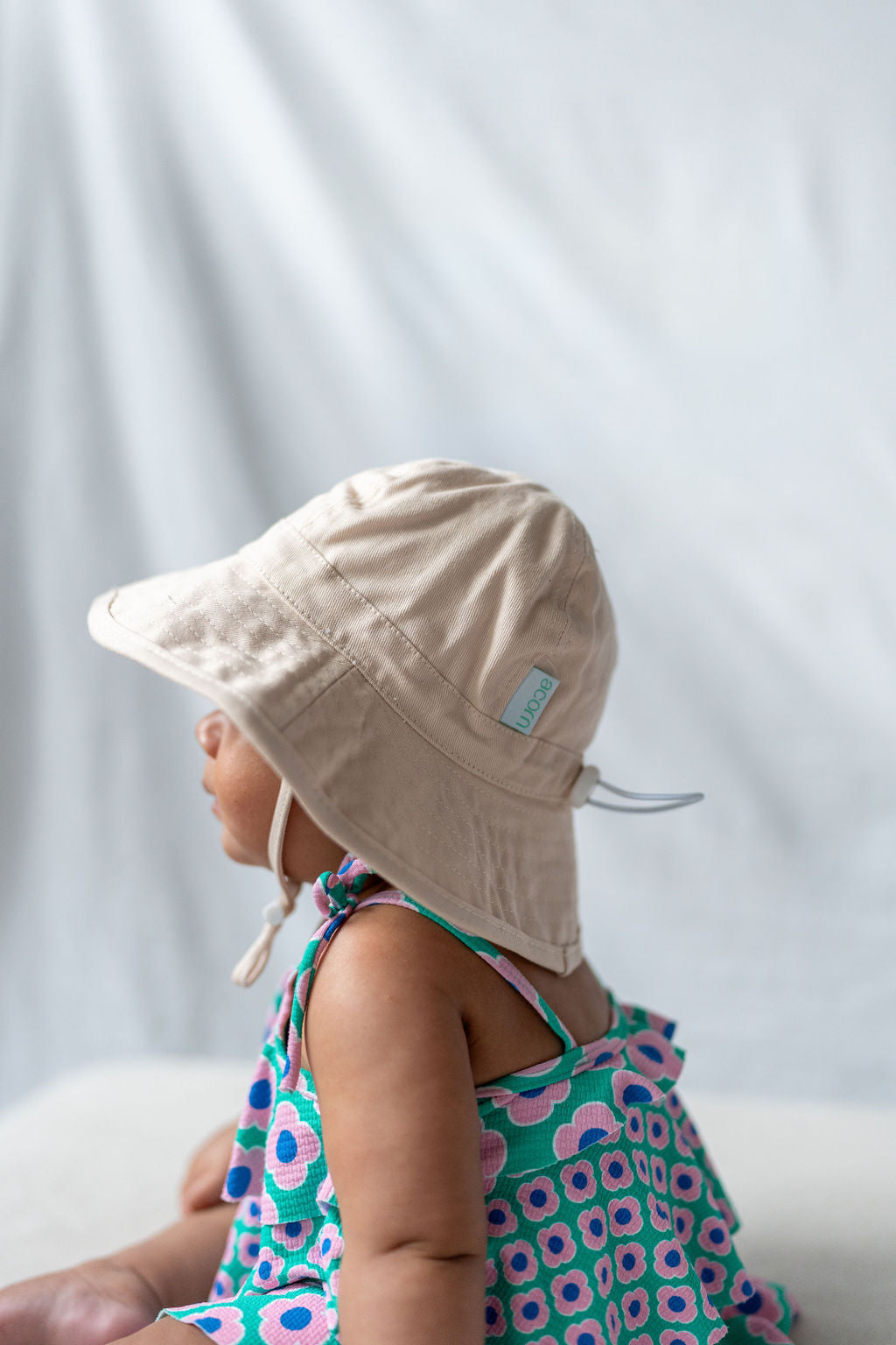 A young child sits sideways facing left, donning the ACORN KIDS Wide Brim Infant Hat Vanilla with a chin strap and UPF50+ sun protection. The floral sleeveless dress made from cotton canvas showcases pink flowers on a turquoise background. The backdrop is plain and softly textured.