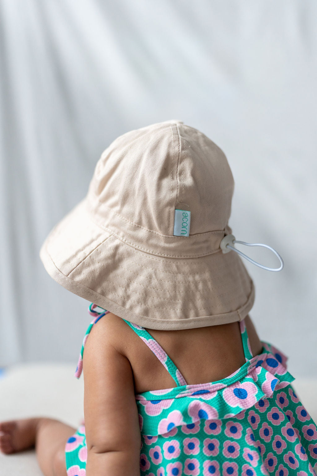 A baby wearing an ACORN KIDS Wide Brim Infant Hat Vanilla with UPF50+ sun protection and a colorful dress adorned with floral patterns is seated facing away from the camera. The scene is softly lit, with a white background providing a gentle, neutral backdrop.