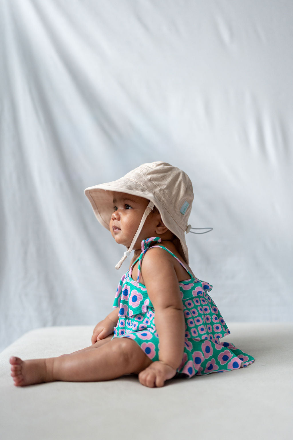 A baby wearing an ACORN KIDS Wide Brim Infant Hat Vanilla made from cotton canvas and a colorful patterned romper sits on a light surface against a plain white background, gazing off to the side.