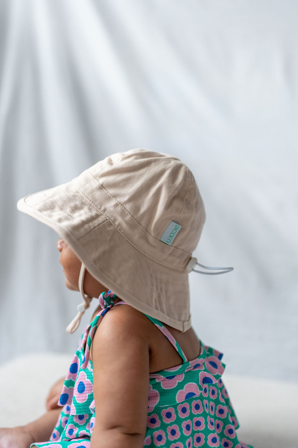 A baby wearing an ACORN KIDS Wide Brim Infant Hat Vanilla with UPF50+ sun protection and a small tag on the side sits facing sideways in a colorful dress featuring a floral pattern against a soft white background.