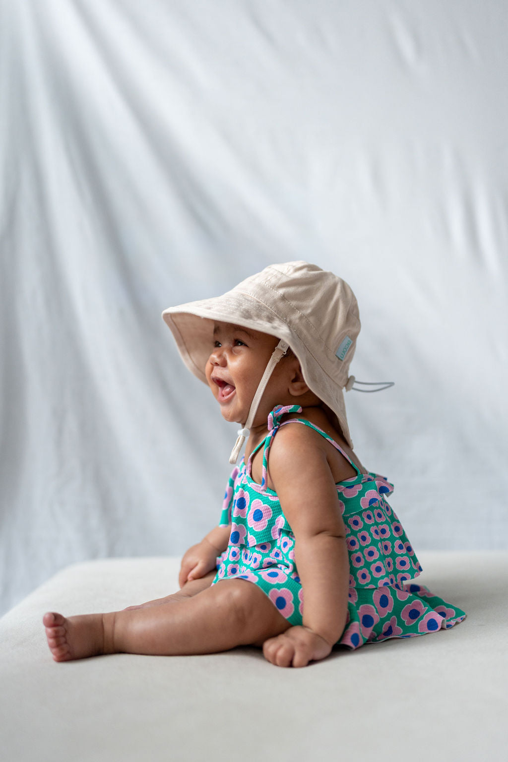 An infant wearing a cute ACORN KIDS Wide Brim Infant Hat in Vanilla, crafted from cotton canvas with UPF50+ sun protection, and a colorful, patterned outfit sits and smiles against a white background.