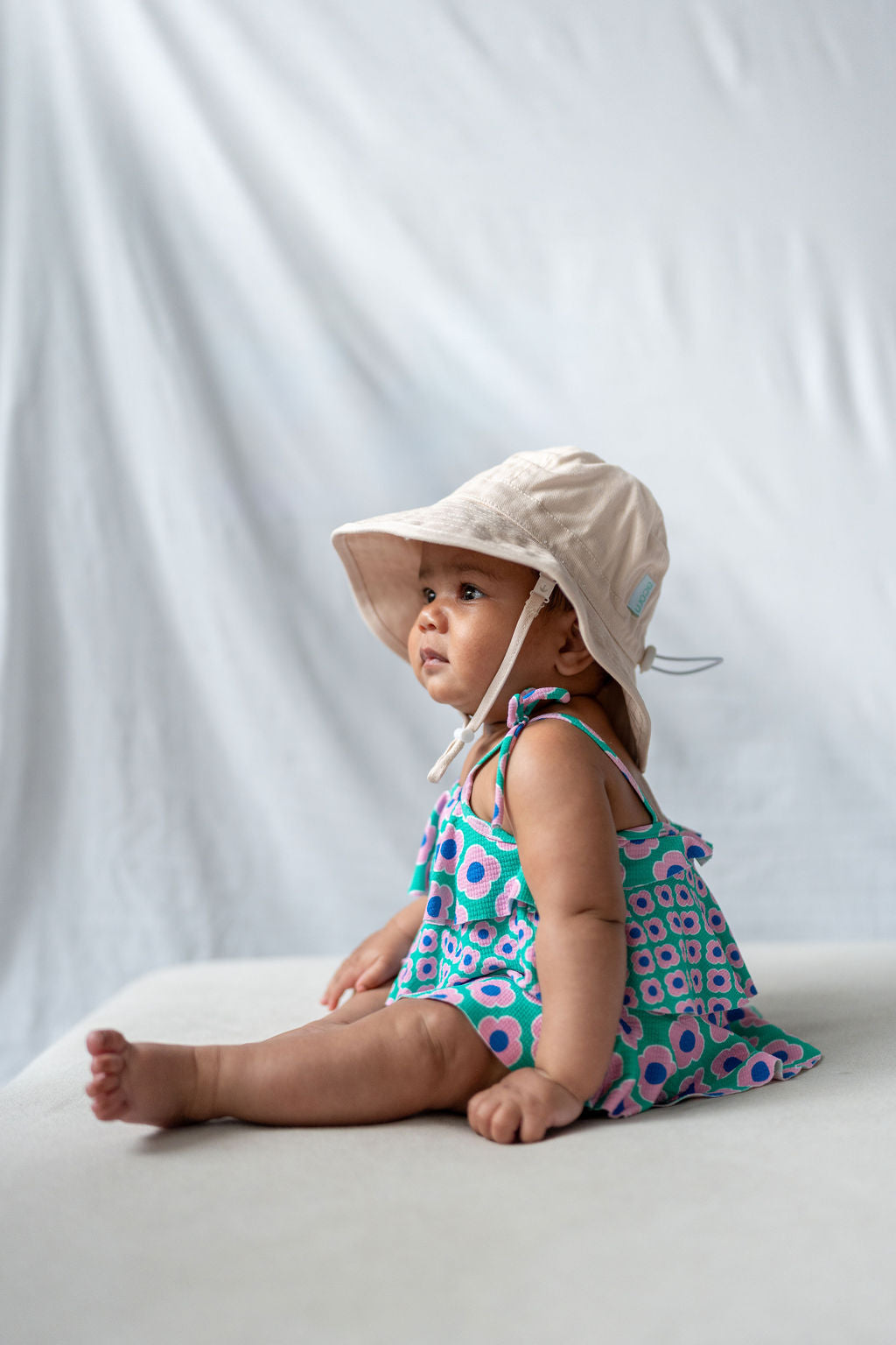 A baby wearing an ACORN KIDS Wide Brim Infant Hat Vanilla with UPF50+ sun protection and a colorful dress decorated with blue, green, and pink floral patterns sits on a soft surface against a plain white backdrop. The baby looks off to the side with a curious expression.