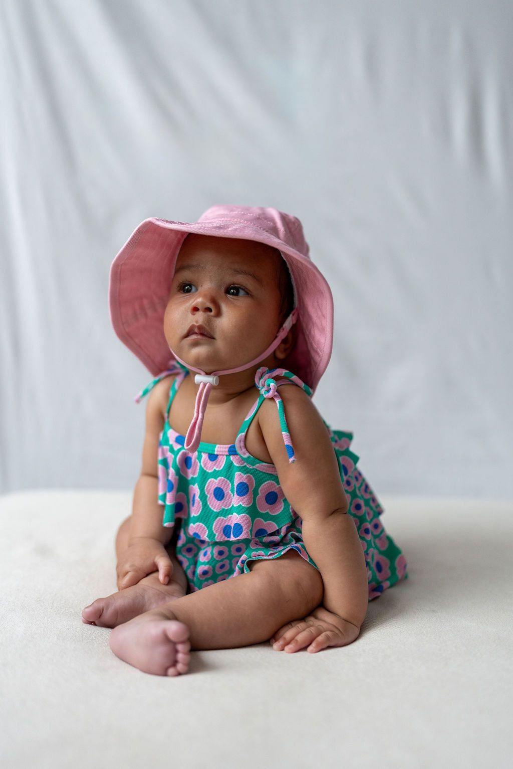 An infant dressed in a blue dress adorned with pink and green floral patterns is seated on a light blanket. The child is also wearing an ACORN KIDS Wide Brim Infant Hat in pink for sun protection, against a light gray backdrop. The baby gazes slightly upward and to the side with a neutral expression.