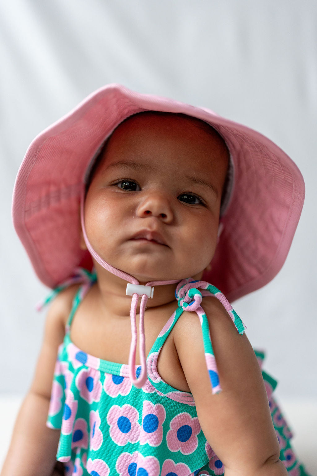 An infant wearing an ACORN KIDS Wide Brim Infant Hat Pink and a colorful dress with a flower pattern looks at the camera. The cotton canvas dress enhances the charm. With a calm expression on their face, the baby enjoys perfect sun protection. The background is a plain, light-colored backdrop.