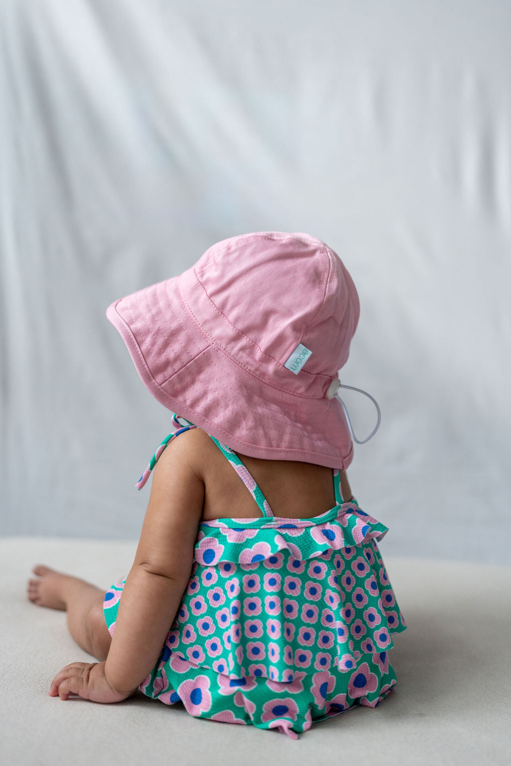 A baby sits facing away from the camera, wearing the ACORN KIDS Wide Brim Infant Hat Pink and a colorful sleeveless dress adorned with green, pink, and blue geometric patterns. The plain, light-colored background underscores the importance of sun protection for little ones.