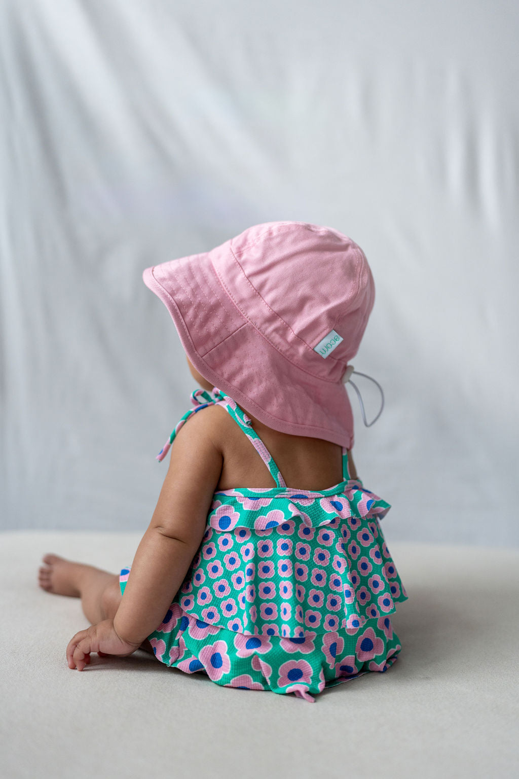 A baby, sitting with their back to the camera, is adorably dressed in the ACORN KIDS Wide Brim Infant Hat Pink for sun protection and a vibrant floral-patterned dress. The soft cotton canvas backdrop enhances the light-hearted charm of the scene.