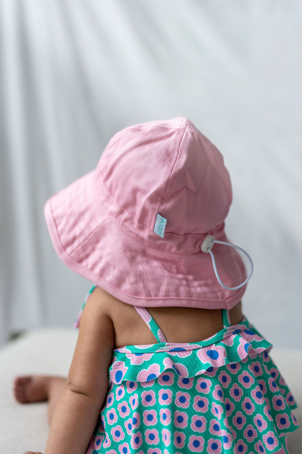 A baby sits with their back to the camera, wearing an ACORN KIDS Wide Brim Infant Hat in pink, made of cotton canvas, and a colorful dress with a blue, green, and pink pattern. The background is a simple white or light-colored fabric, providing perfect sun protection for the little one.