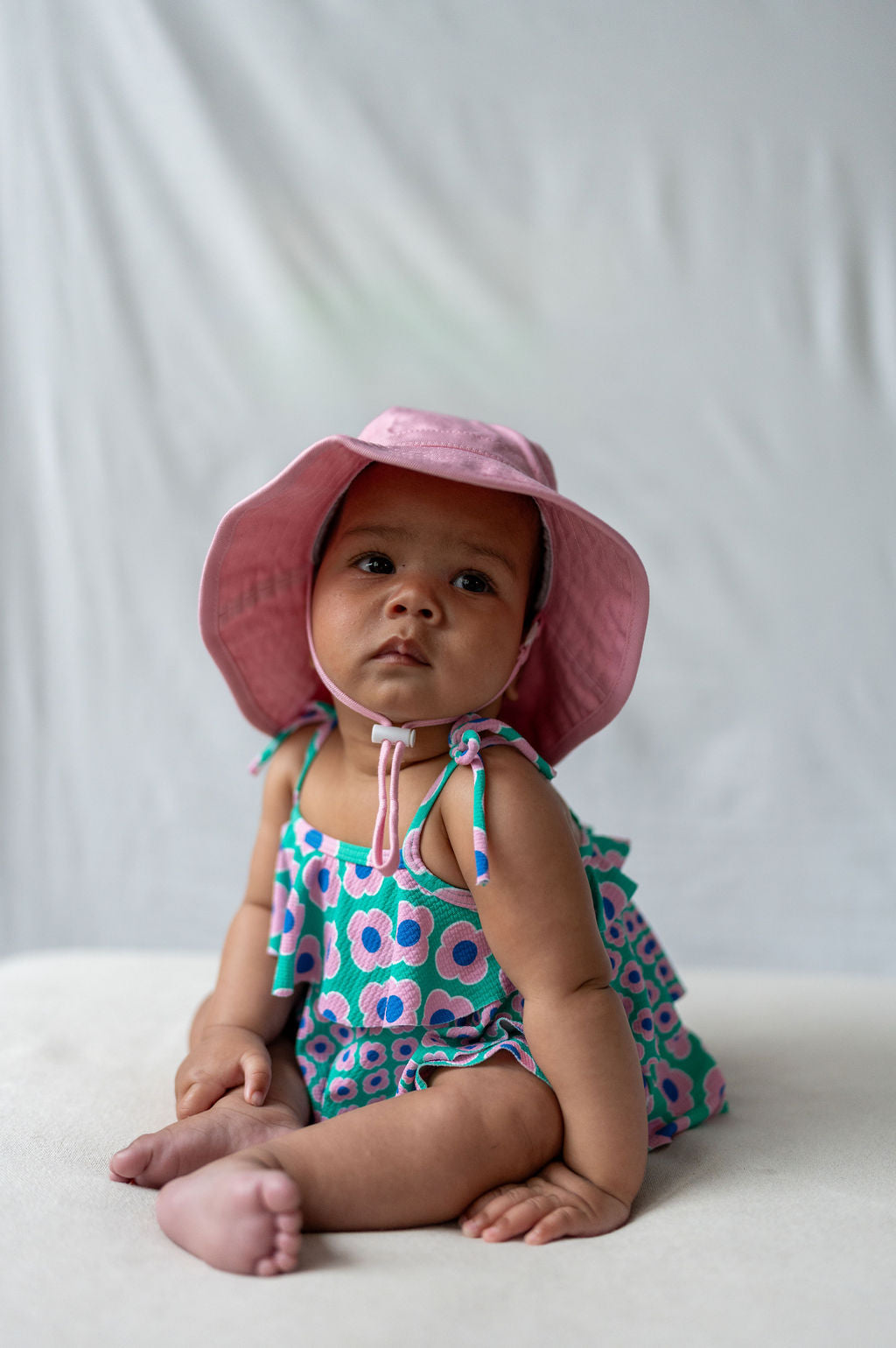 A baby is sitting on a soft surface, wearing a colorful, patterned romper and an ACORN KIDS Wide Brim Infant Hat Pink that offers excellent sun protection with its pink design and secure chin strap. The baby looks slightly to the side against a plain, light gray background.