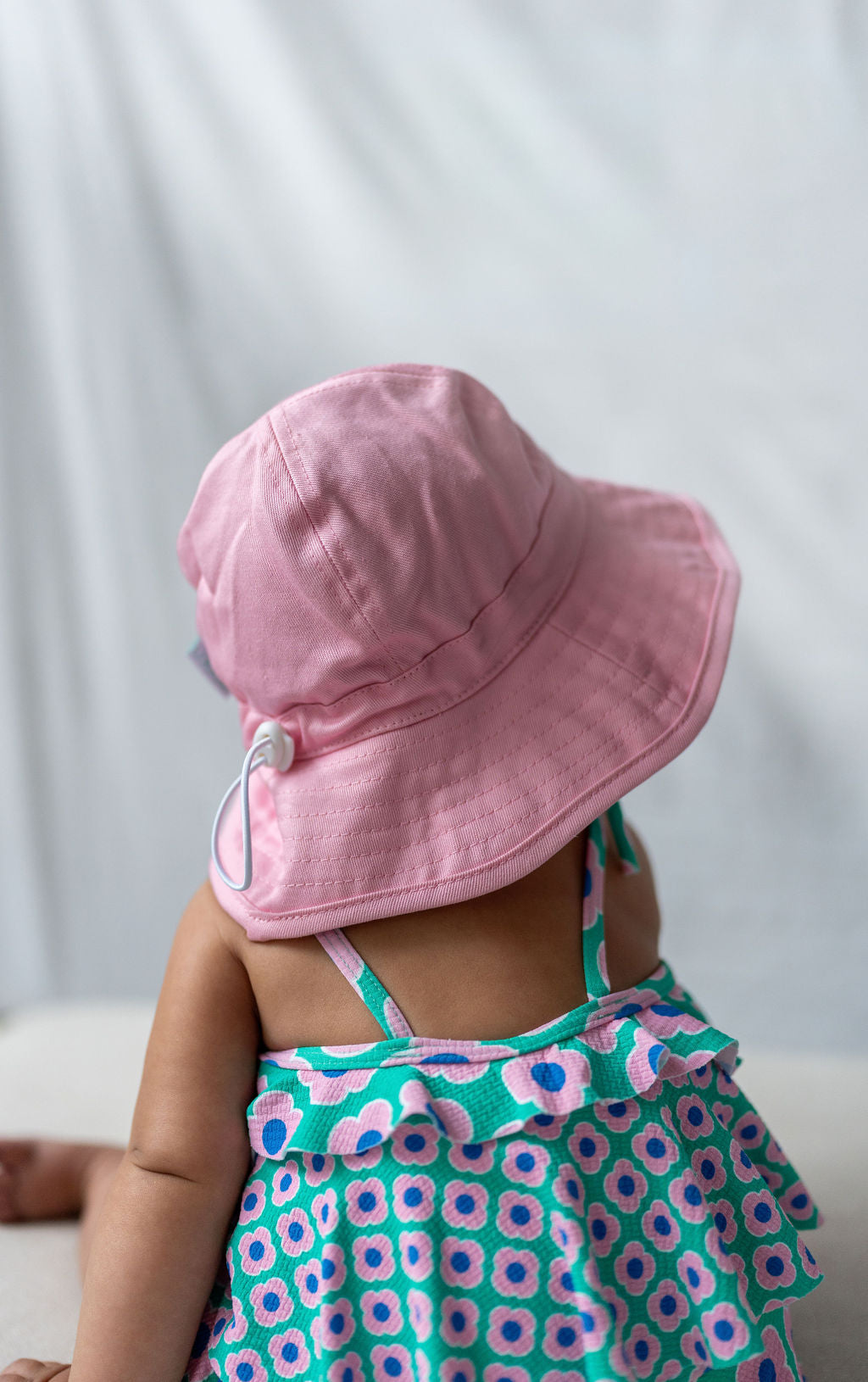 A baby, adorned in an ACORN KIDS Wide Brim Infant Hat Pink with UPF50+ sun protection and a colorful dress featuring a floral pattern, sits facing away from the camera against a plain, light background.