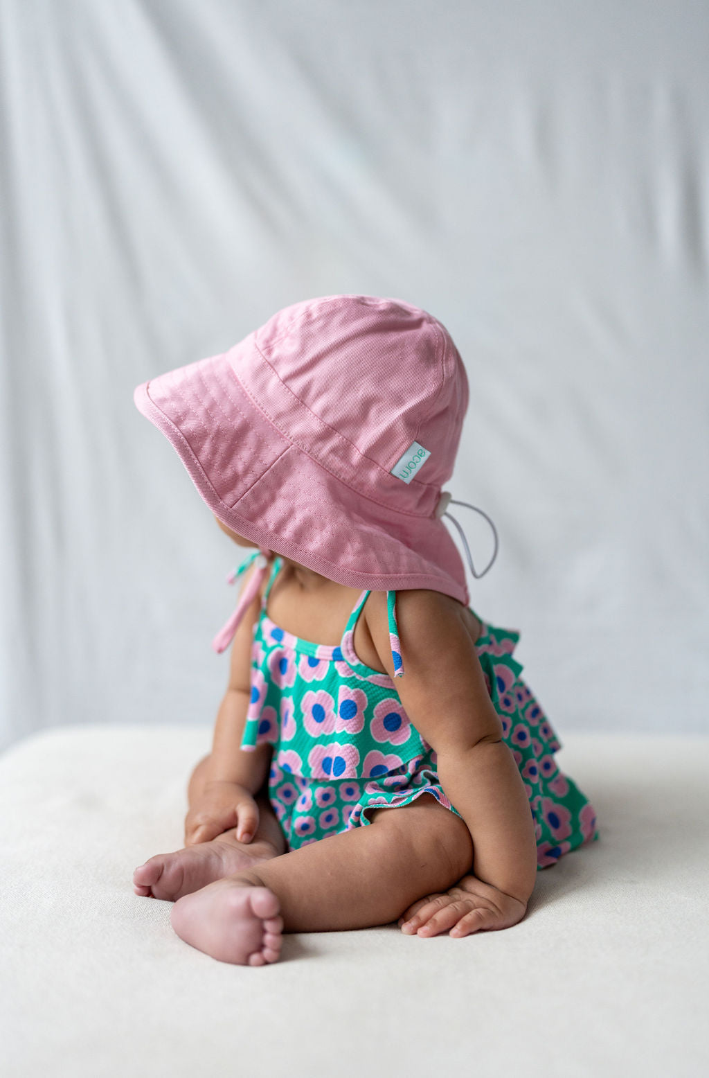 A baby wearing an ACORN KIDS Wide Brim Infant Hat Pink with UPF50+ sun protection sits on a soft surface. Dressed in a colorful floral romper with shades of blue, pink, and green, the baby contrasts softly against the light background, their face turned away from the camera.