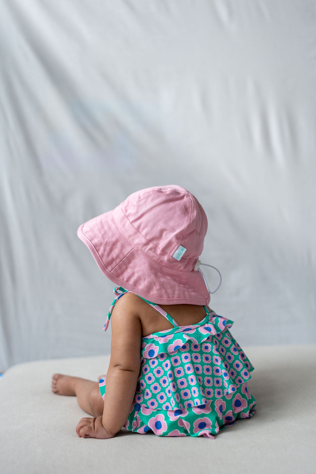 A baby wearing a colorful dress with a geometric pattern and an ACORN KIDS Wide Brim Infant Hat Pink, which offers UPF50+ sun protection, sits facing away on a light cotton canvas surface against a neutral background. The baby is sitting with their legs outstretched, and the soft lighting suggests a calm and serene setting.