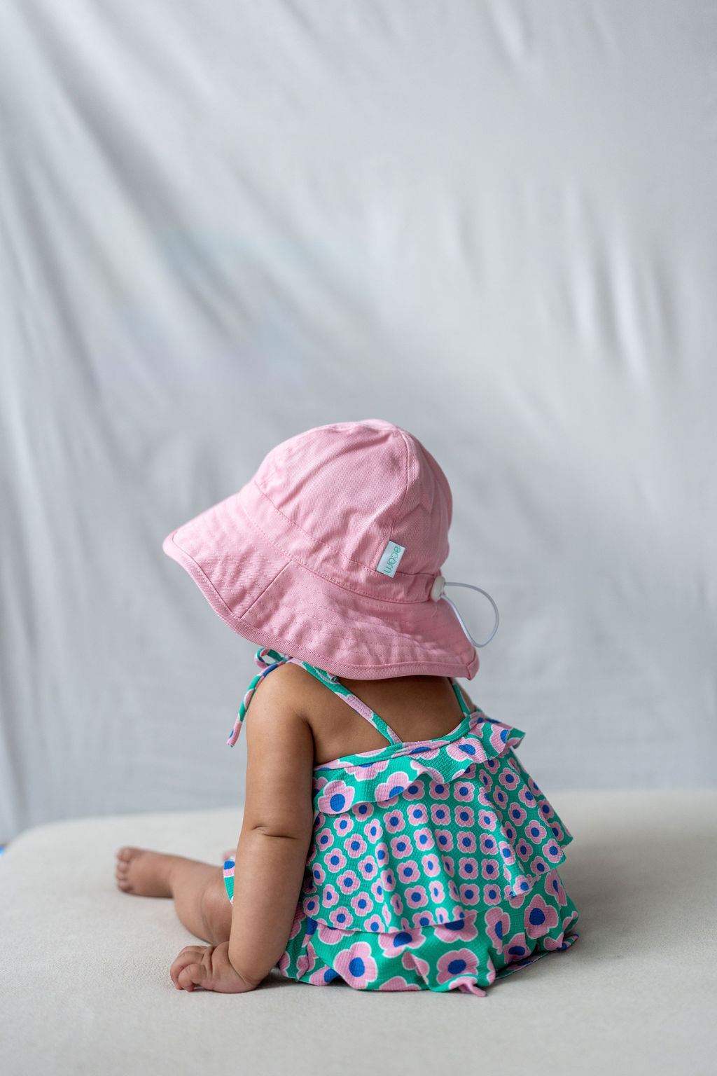 A baby sits facing away from the camera on a light-colored surface, wearing an ACORN KIDS Wide Brim Infant Hat Pink made of cotton canvas that offers UPF50+ sun protection, and a colorful outfit with a green, pink, and blue pattern. The background is a plain, soft white fabric backdrop.