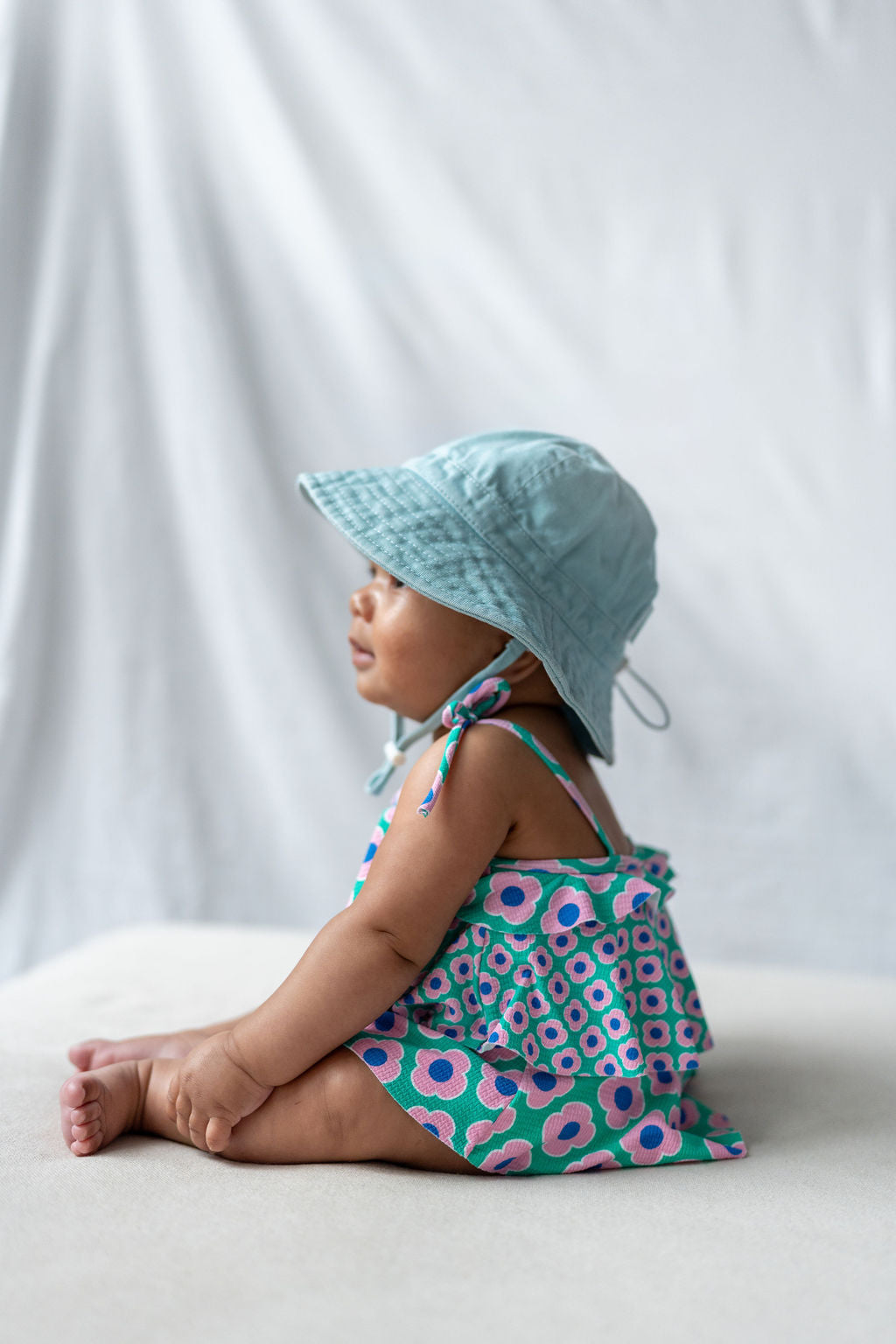 A baby wearing a green, pink, and white floral-patterned romper and an ACORN KIDS Wide Brim Infant Hat Blue with UPF50+ sun protection sits on a white surface with legs outstretched. The background is a soft, neutral-colored fabric. The baby is turned to the side, showing their profile.