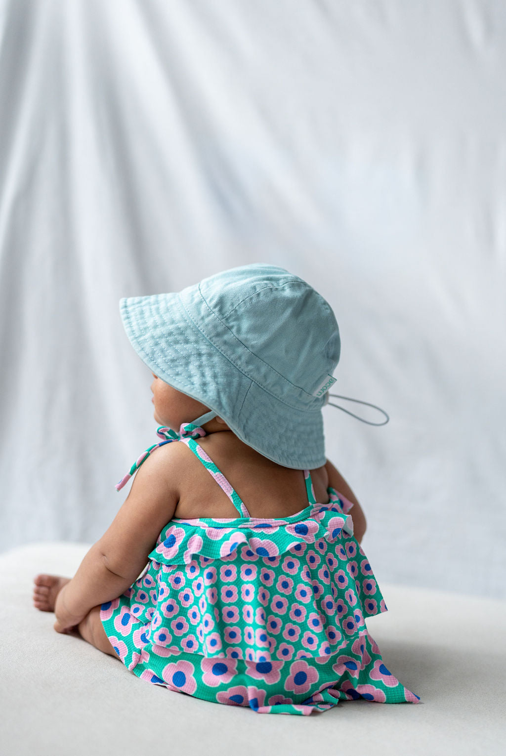 A baby sits with their back to the camera, wearing an ACORN KIDS Wide Brim Infant Hat in blue that offers UPF50+ sun protection and a colorful dress adorned with pink and purple flowers. The background is a white, slightly textured fabric.