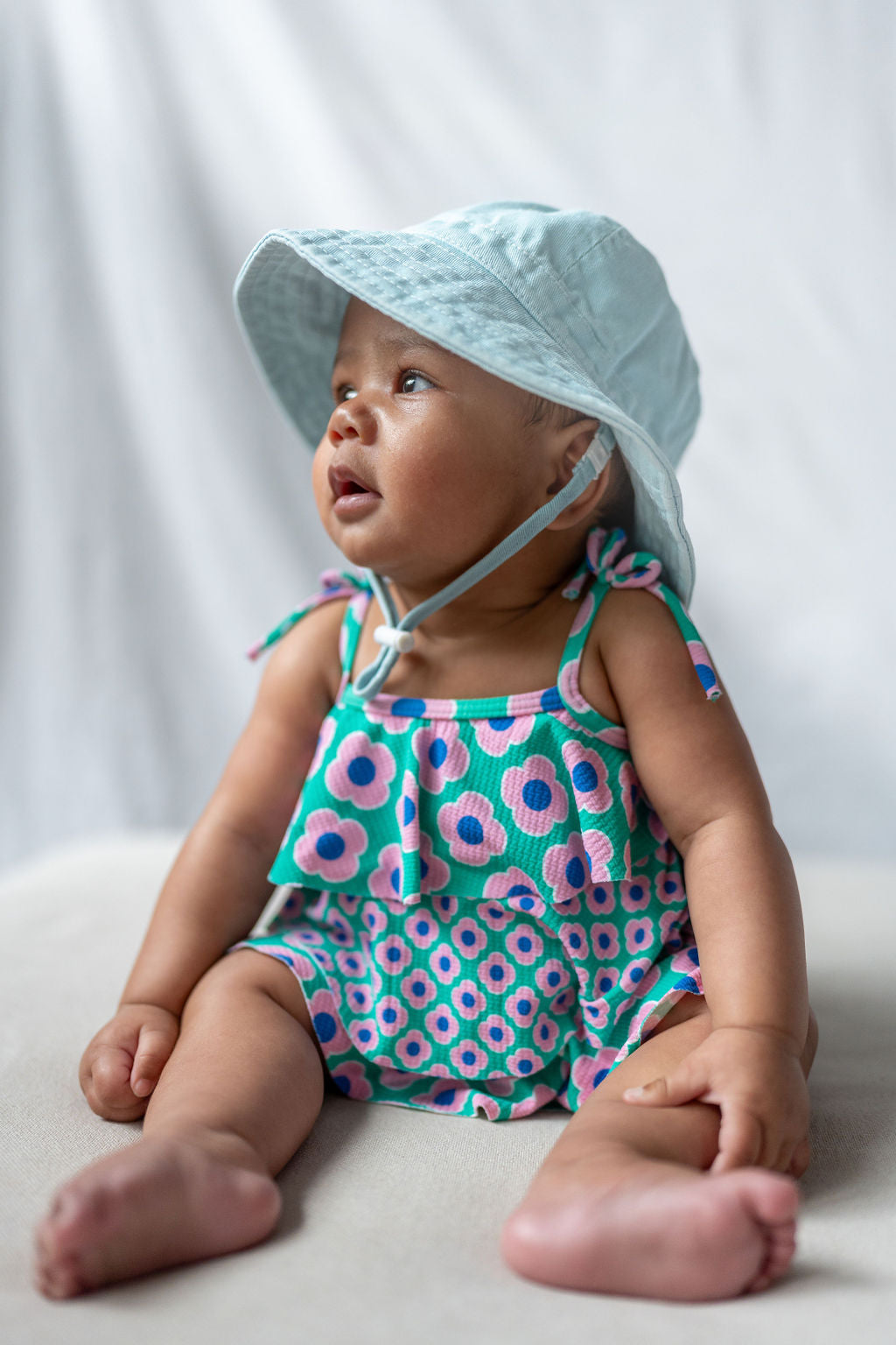 A baby wearing an ACORN KIDS Wide Brim Infant Hat Blue and a floral-patterned outfit sits on a soft surface against a neutral background. The UPF50+ sun protection hat shields the baby's light skin tone as they gaze upward with a curious expression.