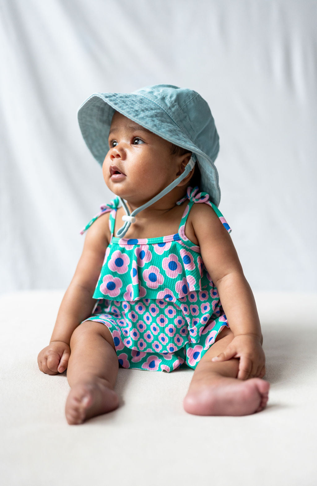 A baby sits on a cotton canvas blanket, wearing an ACORN KIDS Wide Brim Infant Hat Blue with UPF50+ sun protection and a colorful romper with blue, pink, and green patterns. The baby looks up with a curious expression, and the plain, soft background adds focus to the baby.