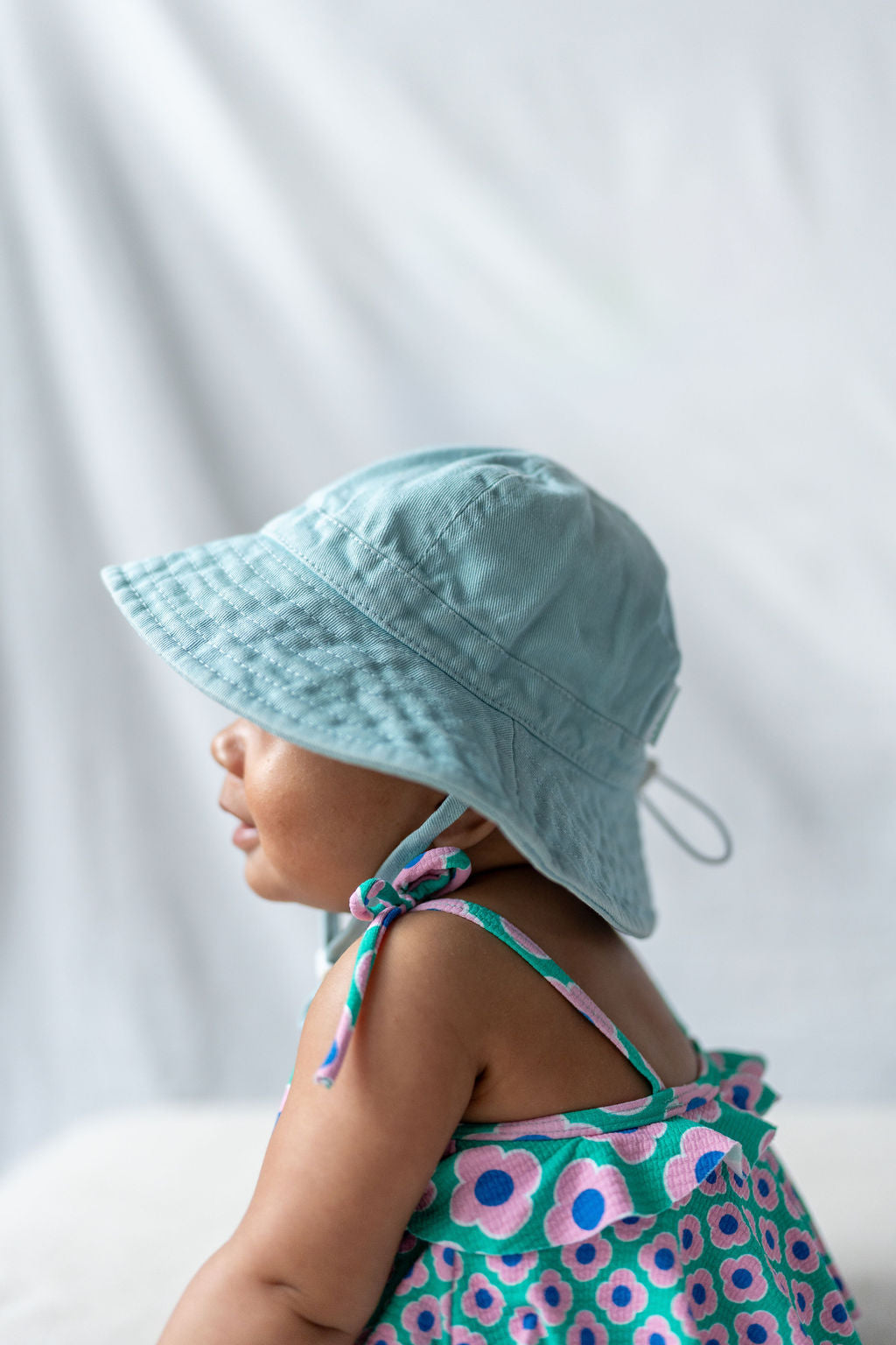 A baby, shown from the side in a colorful floral-patterned outfit, wears the Wide Brim Infant Hat Blue by ACORN KIDS with UPF50+ sun protection. The soft and blurred background makes the child the focal point of the image.