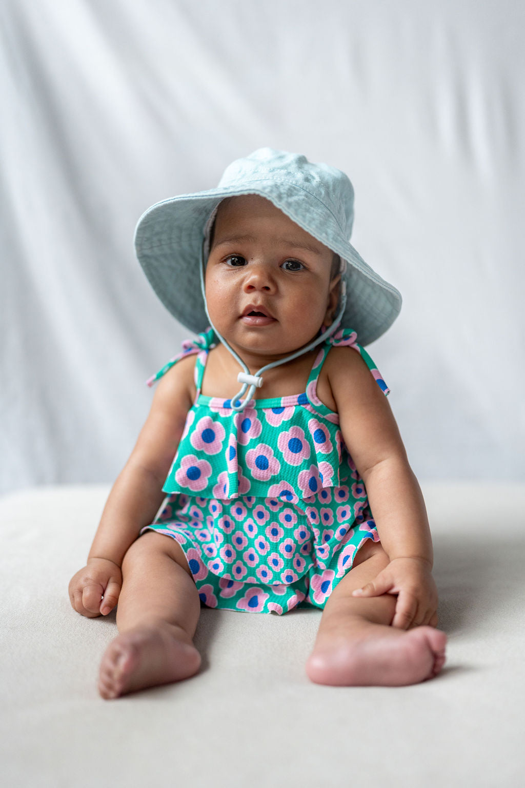 A baby is sitting on a light surface against a plain background, wearing an ACORN KIDS Wide Brim Infant Hat Blue that offers UPF50+ sun protection and a colorful floral romper. The baby's feet are bare, and the expression is neutral, gazing towards the camera.