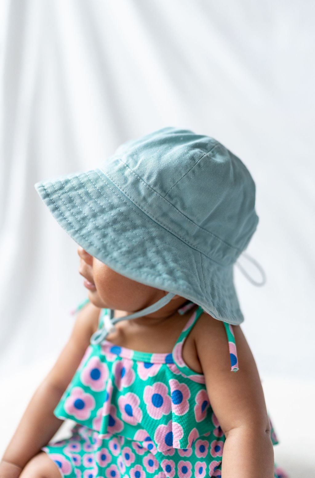 A baby wearing an ACORN KIDS Wide Brim Infant Hat Blue made of cotton canvas with UPF50+ sun protection sits with their head turned to the side. The background is a soft white fabric, creating a gentle and calm setting.