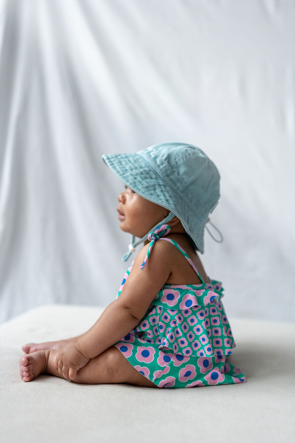 A baby sits on a light surface, turned slightly to the side, wearing a colorful floral dress and an ACORN KIDS Wide Brim Infant Hat Blue offering UPF50+ sun protection. The background is white and simple, drawing attention to the baby's profile.