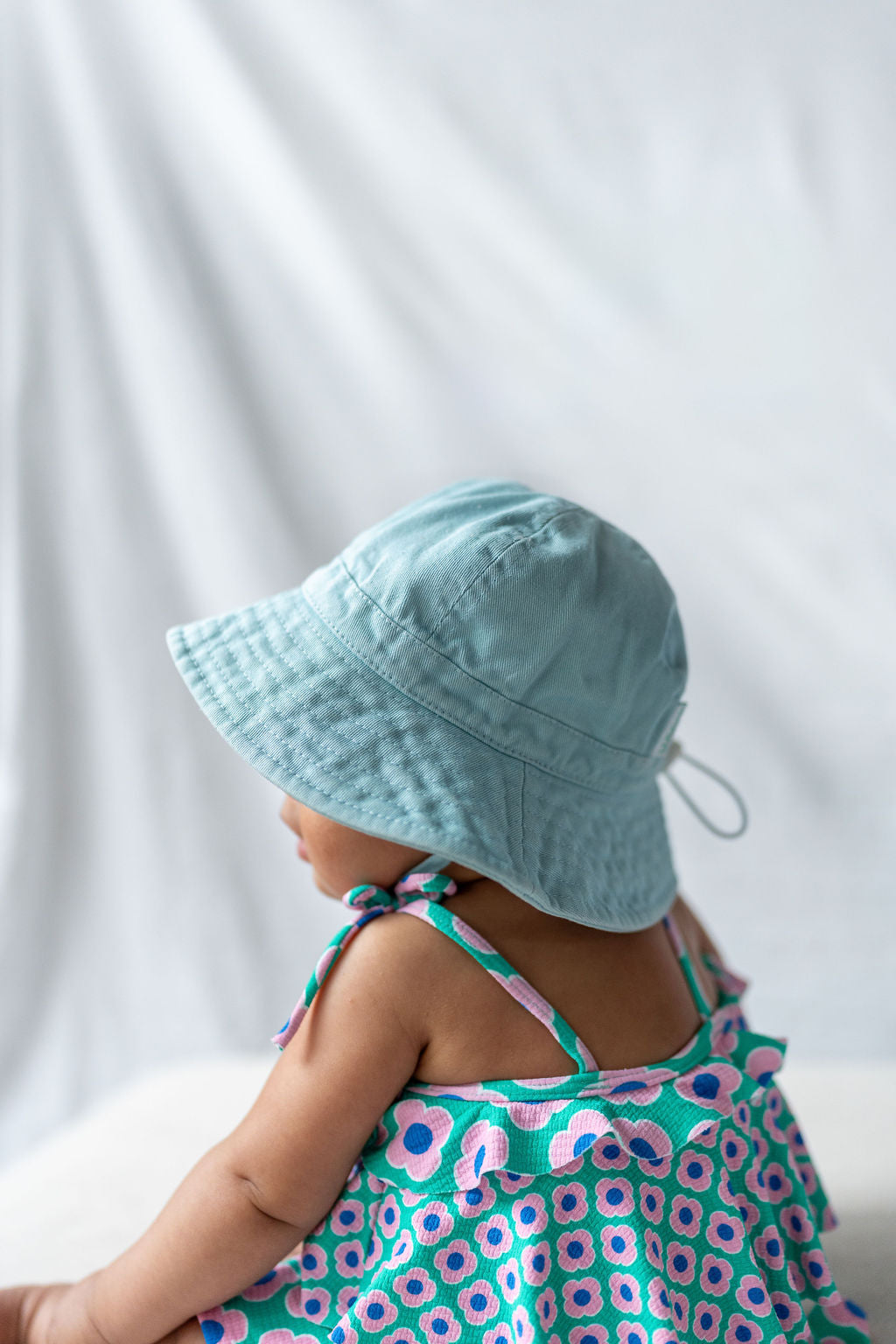 An infant, donning the ACORN KIDS Wide Brim Infant Hat in Blue for sun protection, sits facing sideways against a light, blurred background. Adorned in a vibrant sleeveless dress with a floral pattern showcasing blue, pink, and green tones, the baby's upper body is displayed while their skin tone is visible.