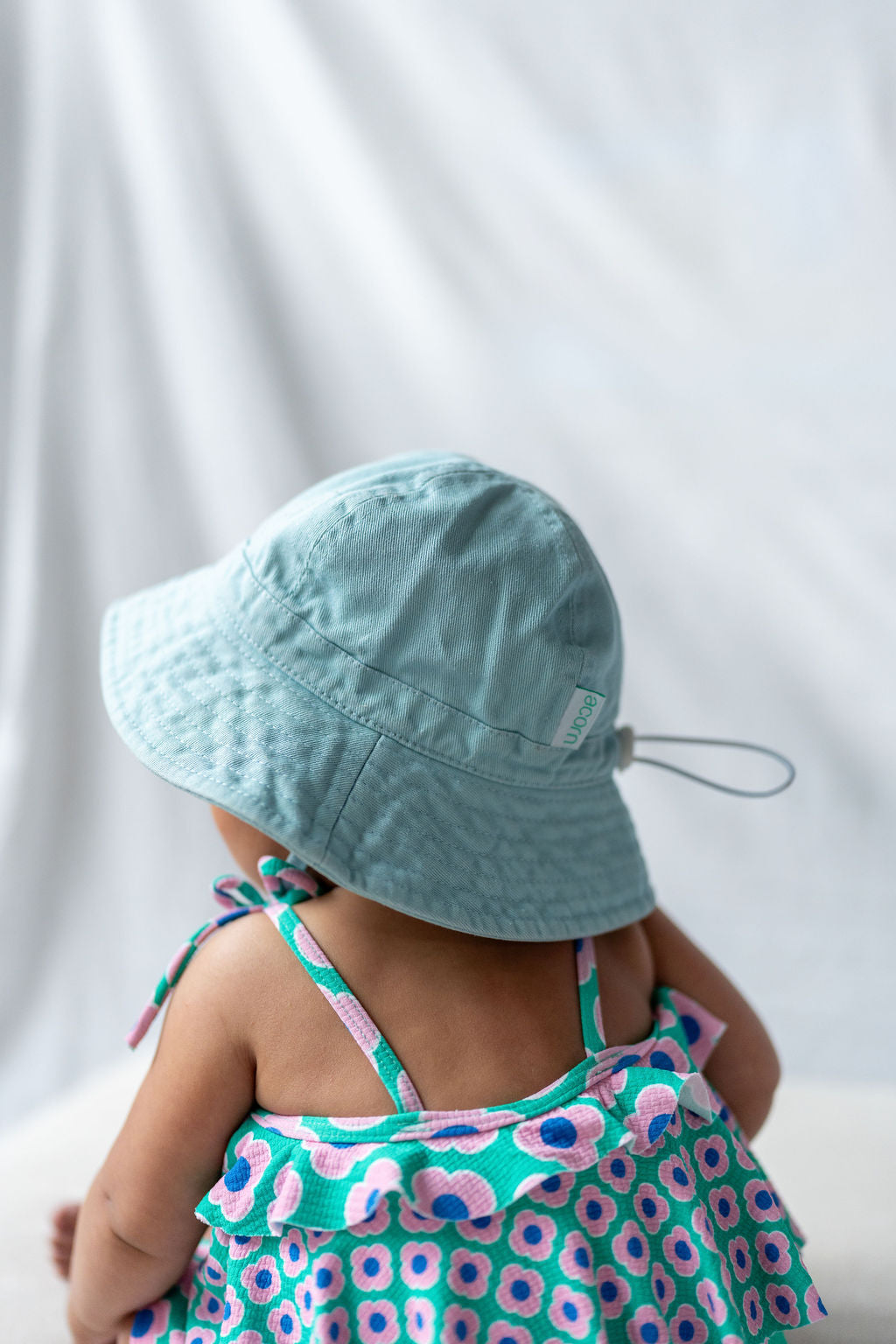 A baby wearing an ACORN KIDS Wide Brim Infant Hat Blue sits with their back to the camera. The hat provides essential sun protection, while the baby is dressed in a colorful, floral-patterned cotton canvas top with pink and blue hues against a soft, light-colored background.