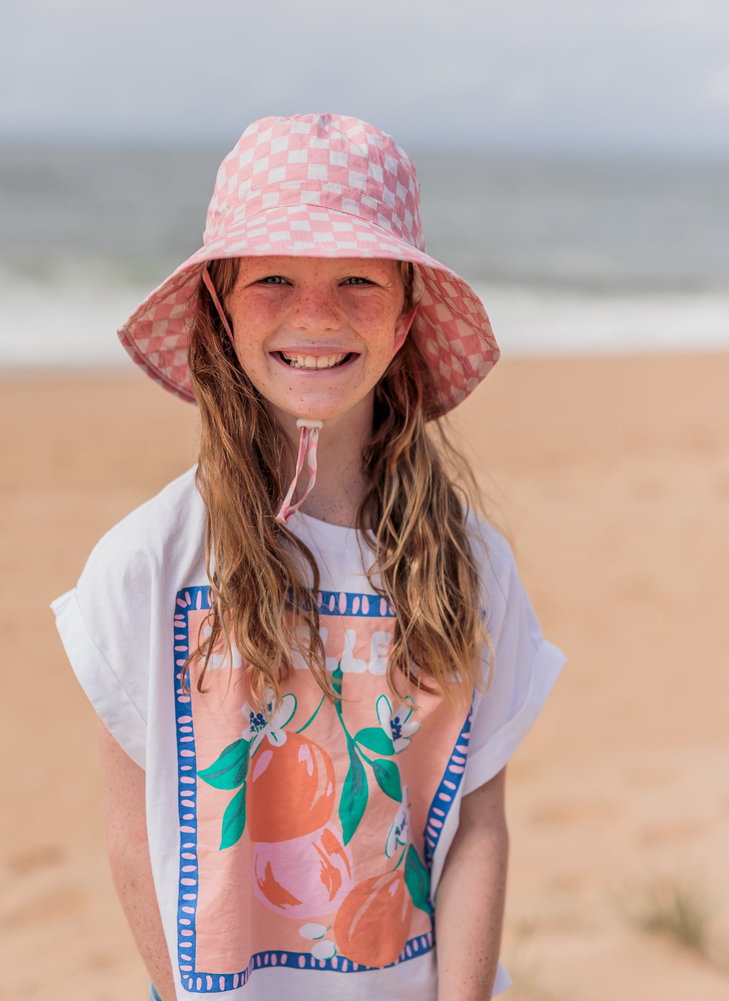 A young girl with long hair smiles on a beach, wearing the ACORN KIDS Pink Checks Broad Brim Bucket Hat for sun protection. Her white shirt is adorned with colorful fruit graphics, while the ocean and sand create the perfect backdrop.