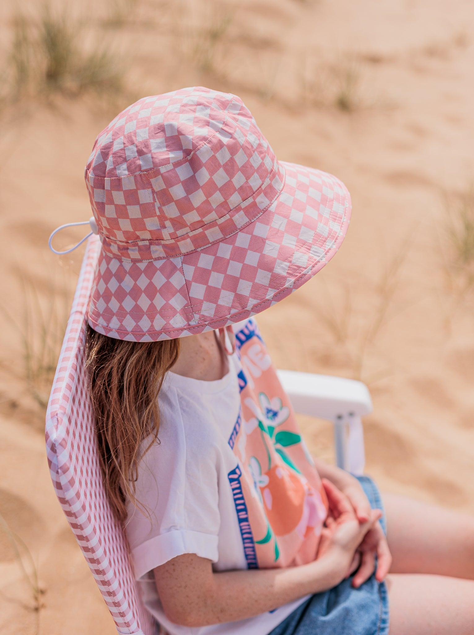 A person wearing an ACORN KIDS Pink Checks Broad Brim Bucket Hat sits on a matching chair at the beach. They don a white t-shirt with a colorful graphic and a denim skirt, embracing sun protection in style against the sandy backdrop with sparse grass.